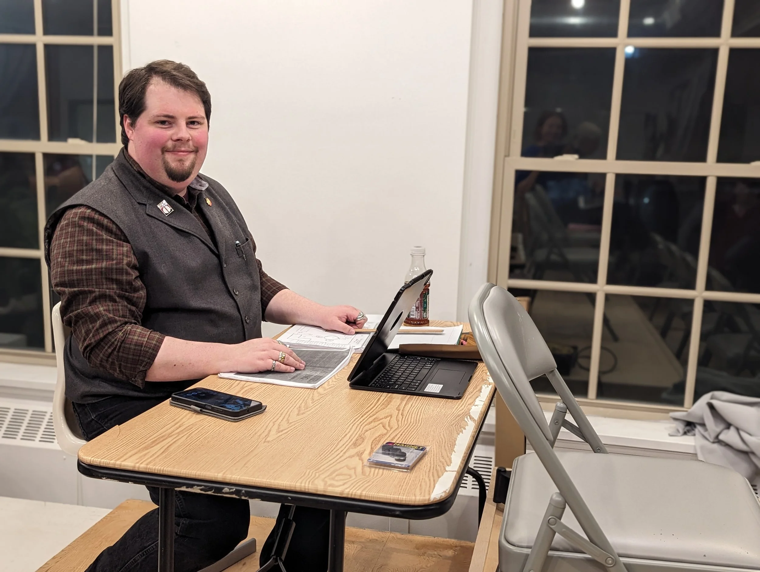 A man with dark hair and a goatee sitting at a wooden table with a laptop, a smartphone, a pen, and a glass soda bottle, smiling at the camera inside a room with large windows.
