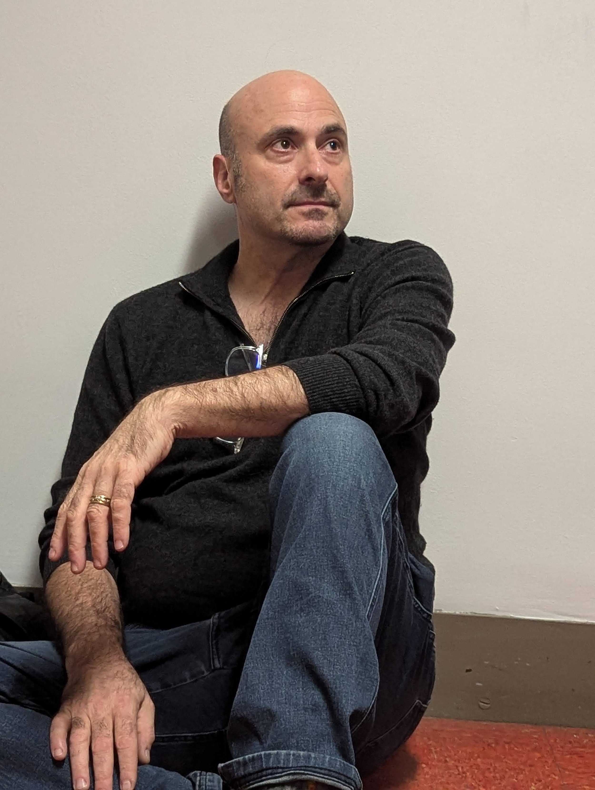 A bald man with a goatee, wearing a black zip-up sweater and blue jeans, sitting on the floor with his knees up, resting against a plain wall, looking to his left.