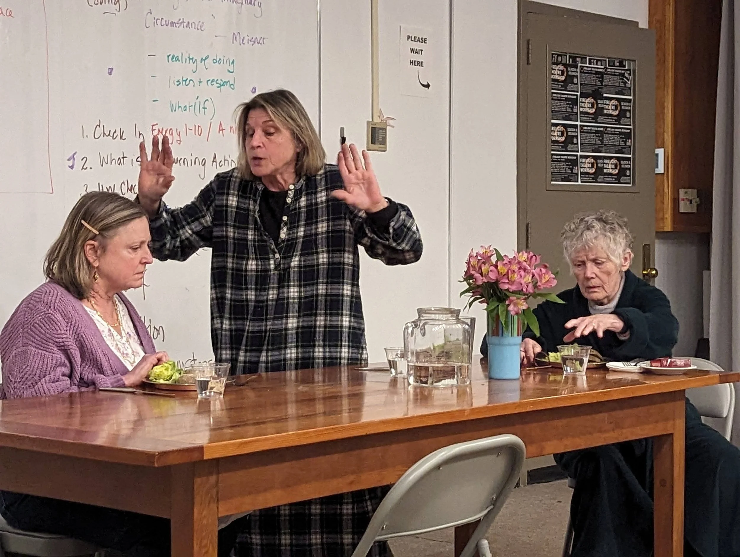 An indoor scene with three women around a wooden table, one standing and talking to the two women seated. One seated woman has a pink cardigan and the other has curly gray hair. There are pink flowers in a blue vase on the table, and a whiteboard wit