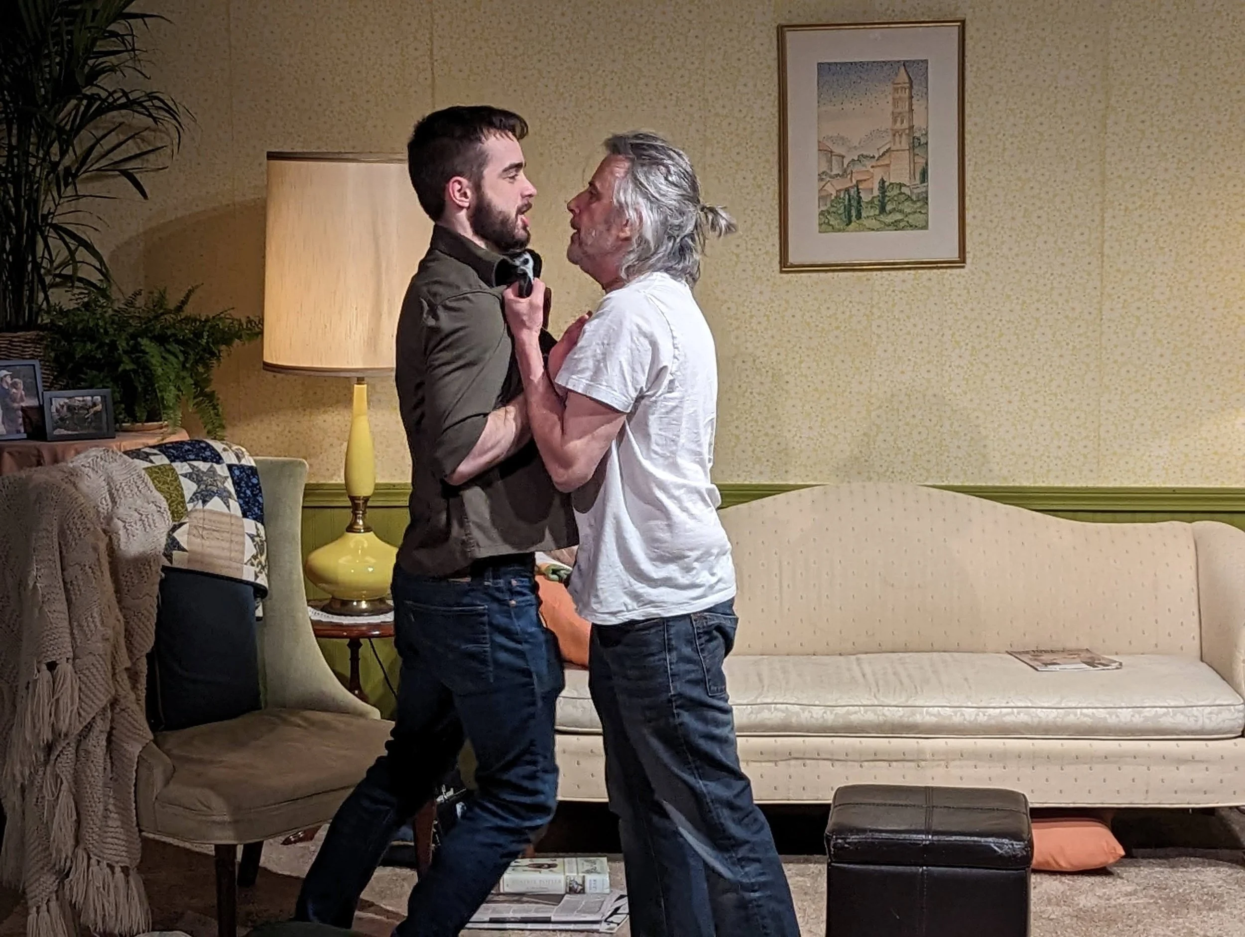 Two men engaged in a heated argument inside a living room. One man, with grey hair and a ponytail, is grabbing the collar of the other man, who has dark hair and a beard.