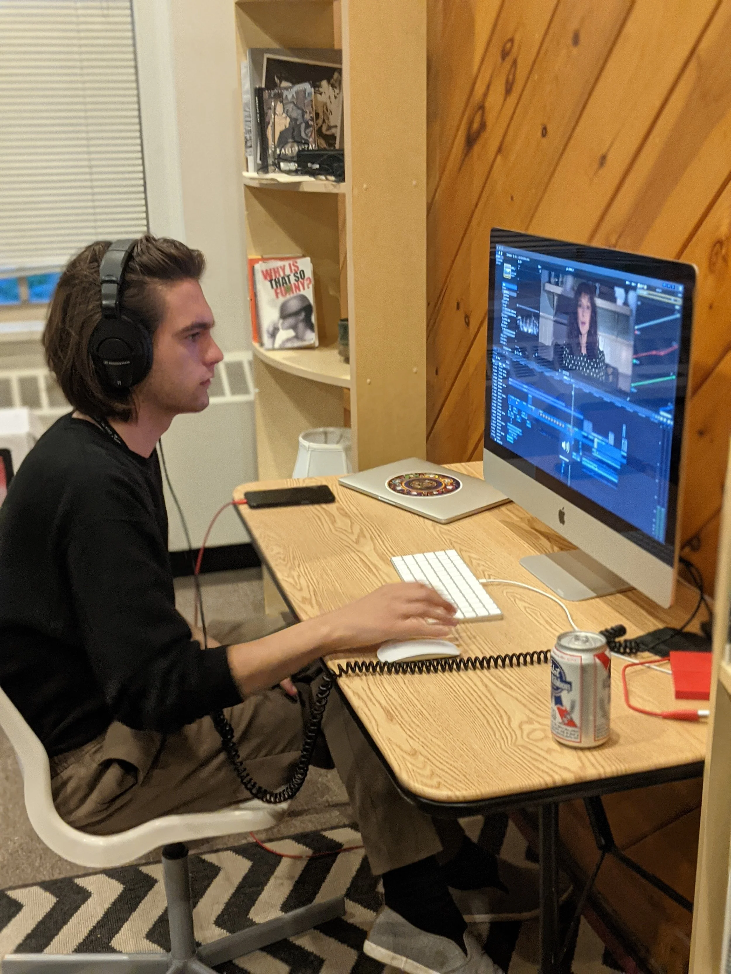 A young man with headphones working on video editing at a desk, with a computer monitor displaying a video editing software and a woman on the screen, near a wooden wall.