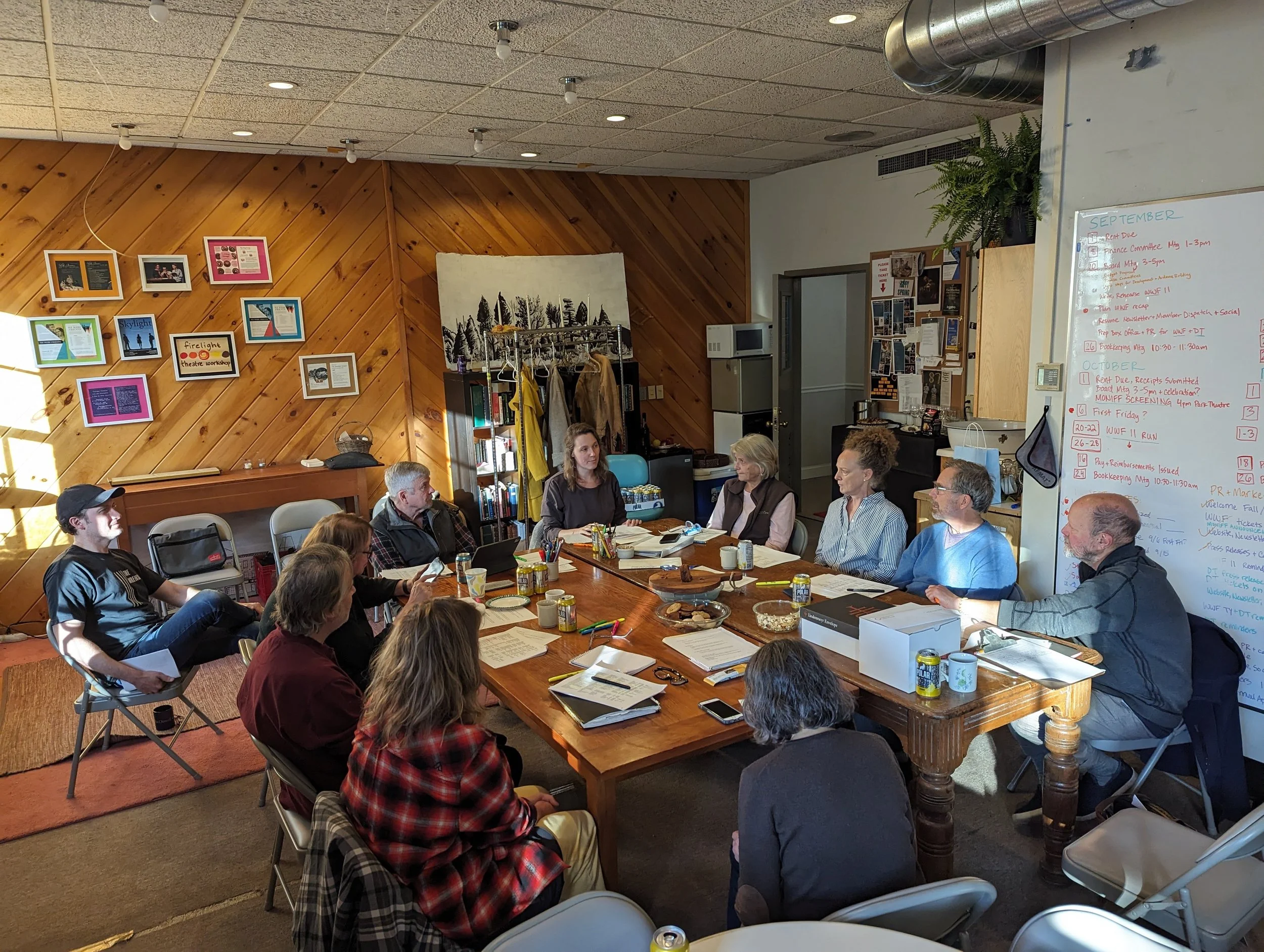 The Firelight Board of Directors sitting around a large wooden table in a meeting room, with a whiteboard on the right side filled with notes and a bulletin board with photos and papers on the back wall.