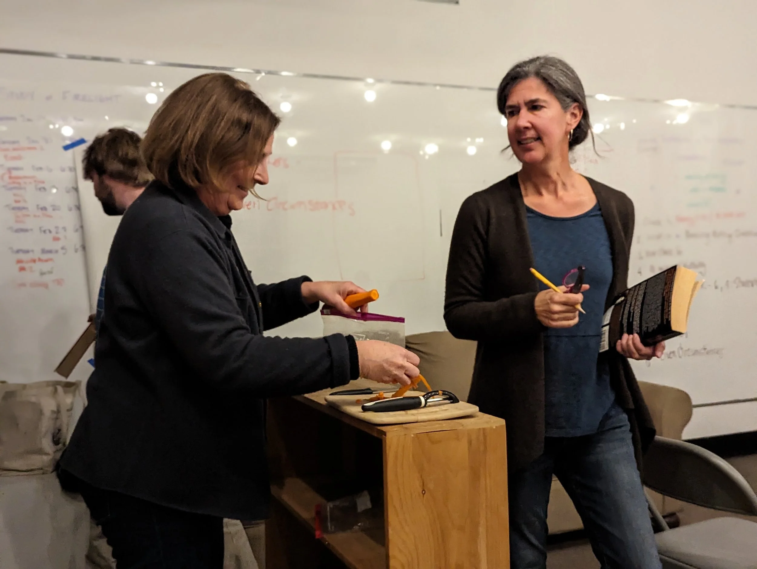 Two women in conversation in a room with a whiteboard in the background. One woman is preparing food or some items on a small wooden table, the other is holding a book and a pen.