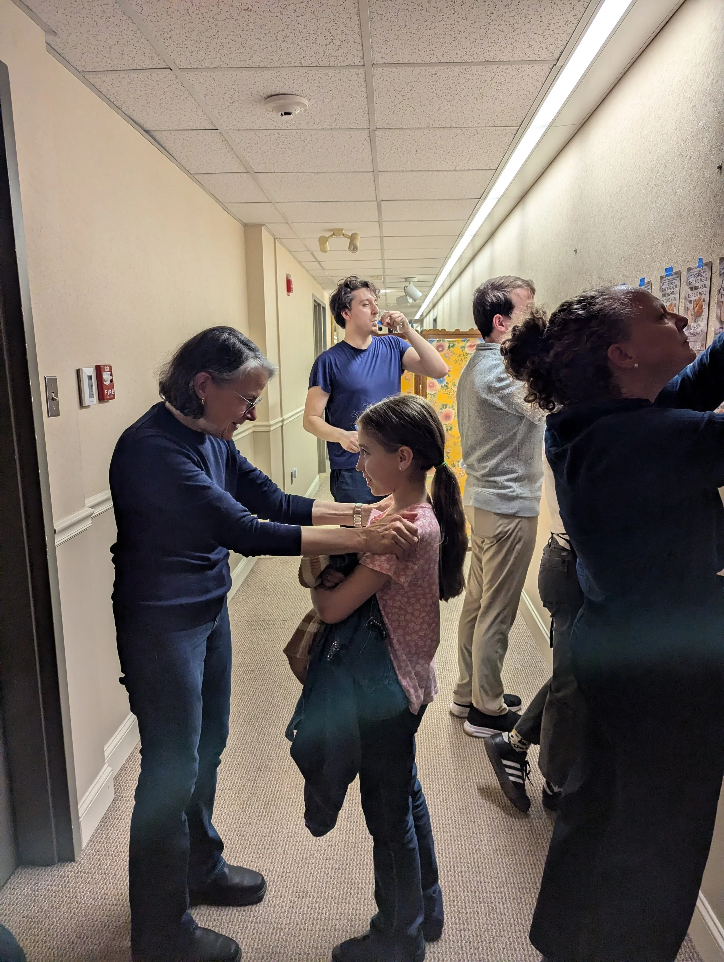 A group of people standing in a line, with a woman helping a young girl with a pin or badge, in an indoor setting. Several people are looking at posters or notices on the wall.