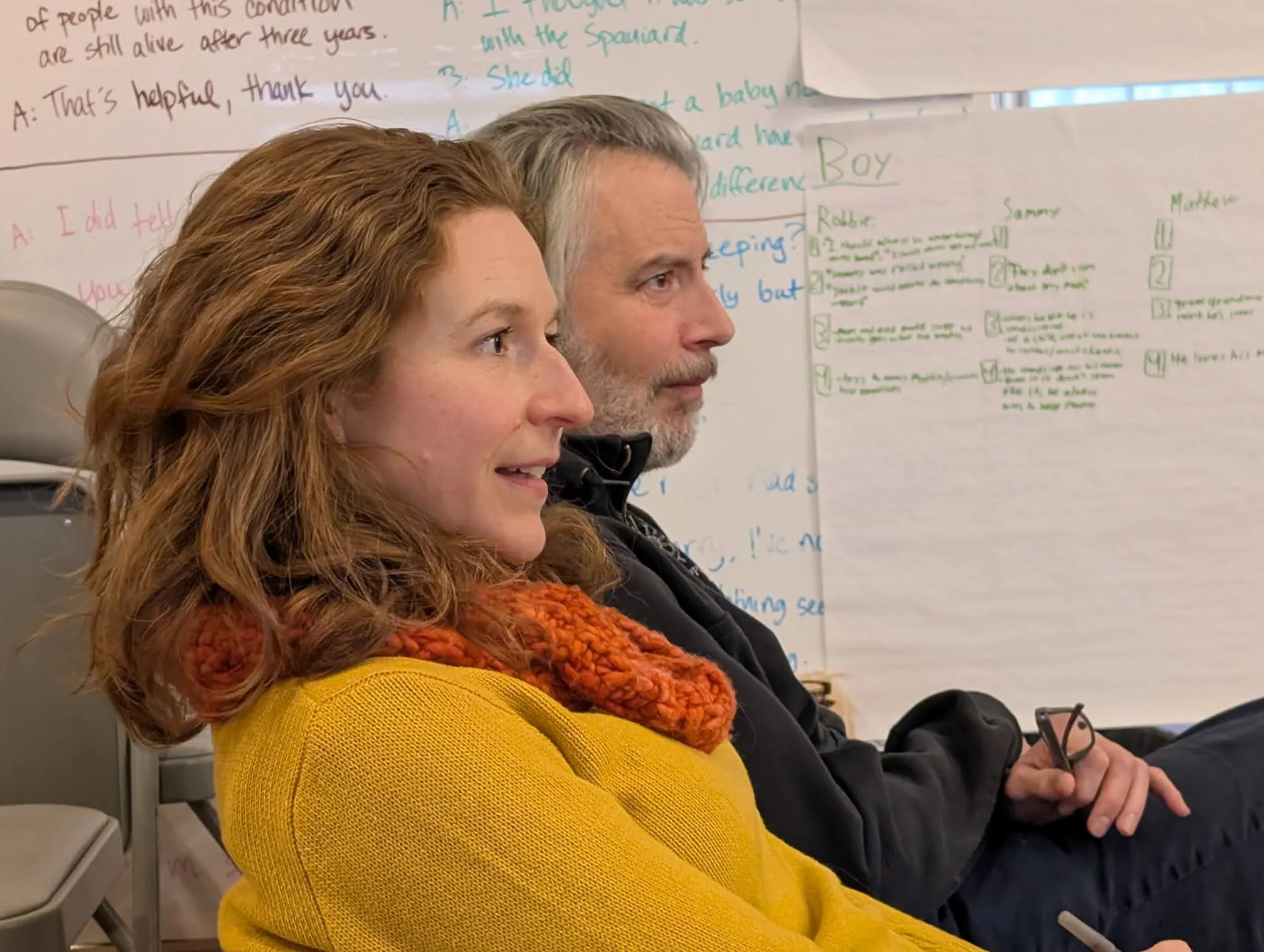 Co-Founders of Firelight, Nora Fiffer and Jason Lambert, sitting in profile, a woman with reddish curly hair wearing a yellow sweater and an orange scarf, and a bearded man with gray hair wearing a dark jacket, watching rehearsal in a studio.