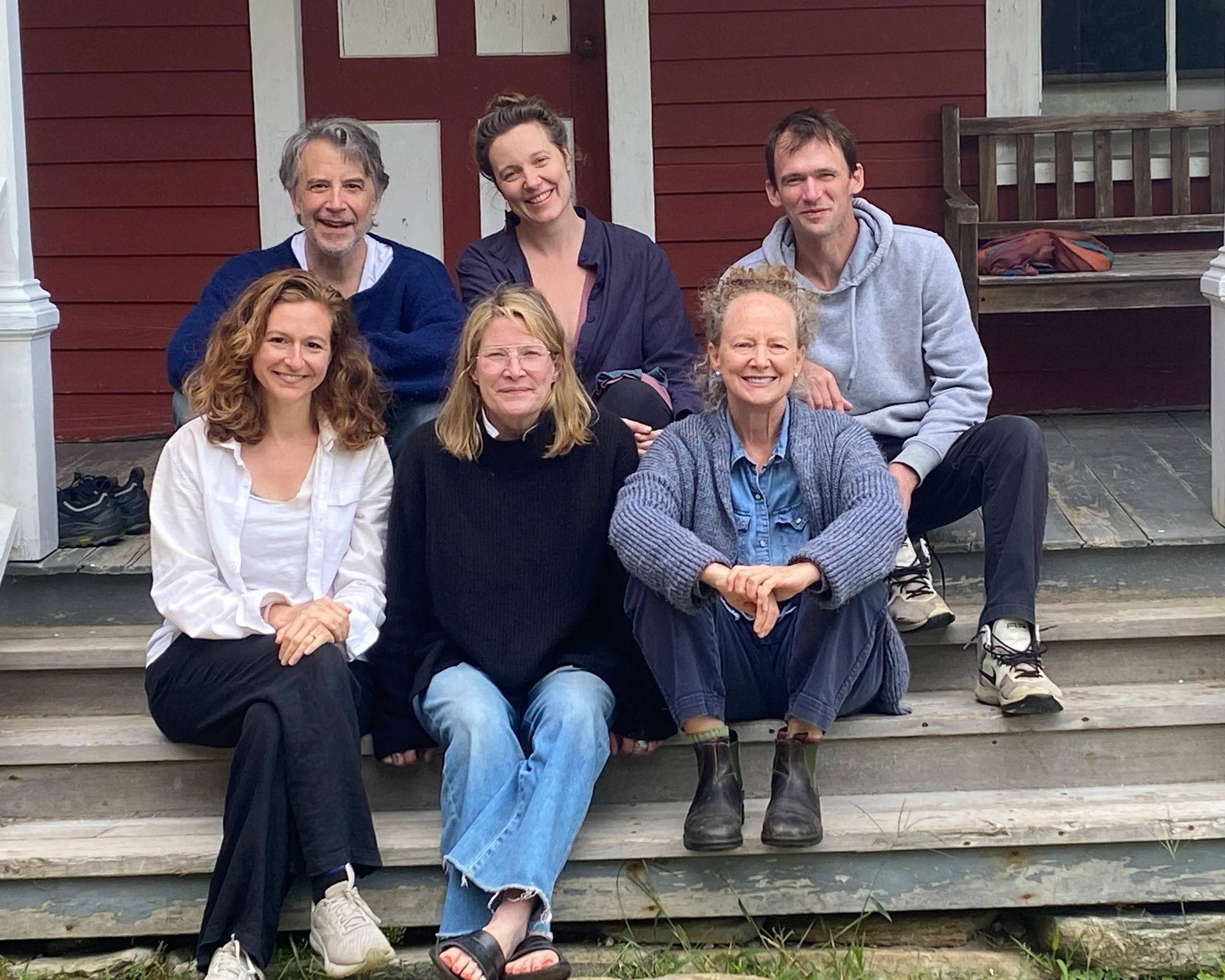 The Firelight Theatre Workshop core artists sitting on front steps of a red house, smiling at the camera. Nora Fiffer, Jason Lambert, Jazimina Creamer-MacNeil, Laura Carden, Sarah Sandback, and Henry Walters.