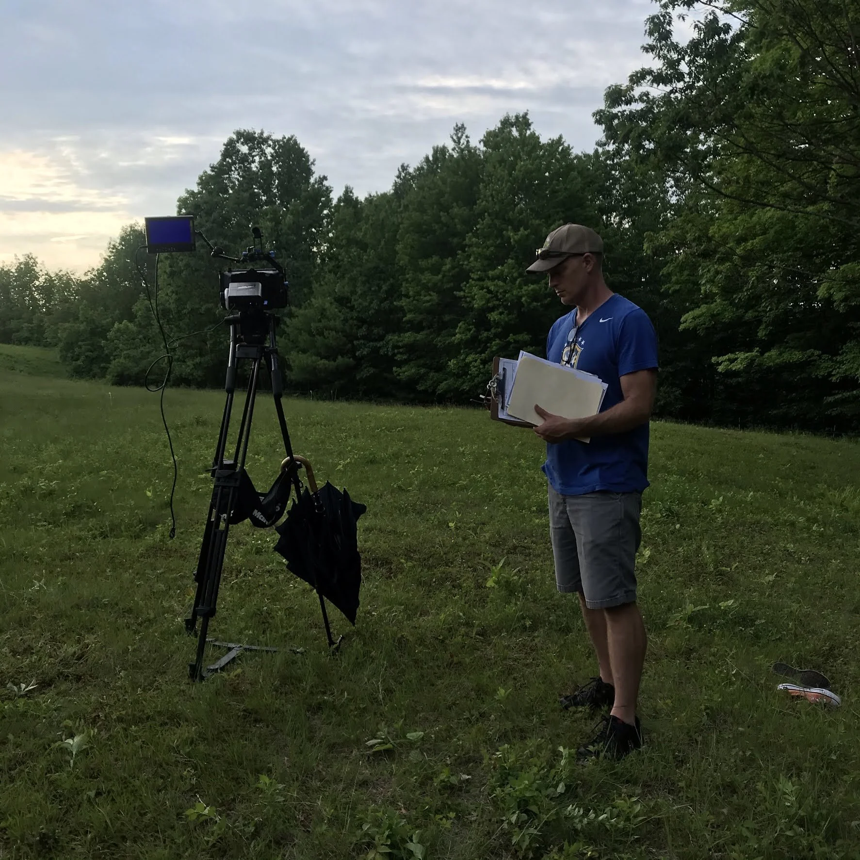 A man in a blue t-shirt, gray shorts, and a beige cap holding a folder or clipboard, standing outdoors next to a video camera on a tripod in a grassy area with trees in the background.