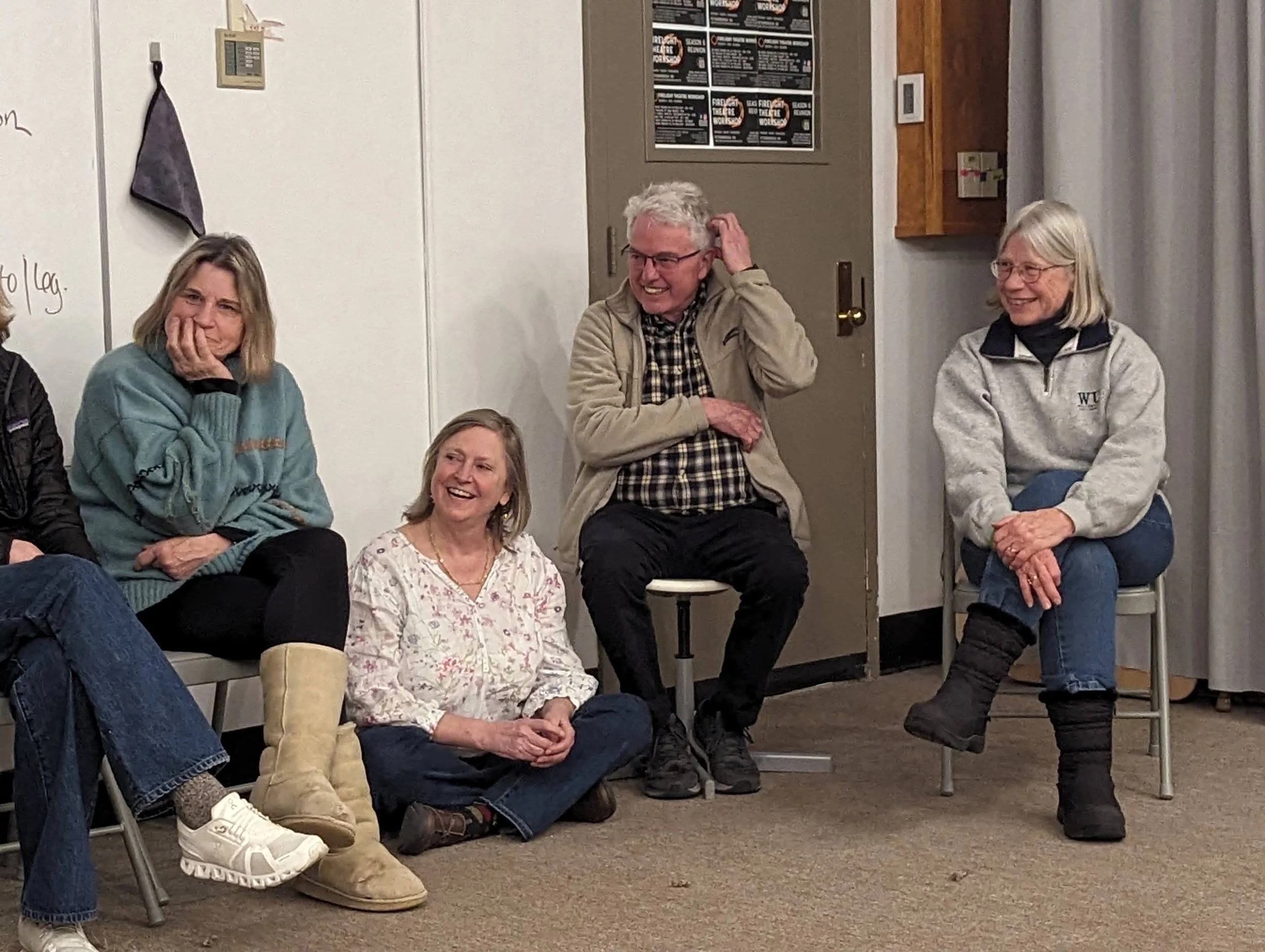 Group of five adults sitting and standing in a room, engaged in conversation and laughter. Four women and one man, one woman is sitting on the floor with crossed legs, the others on chairs.