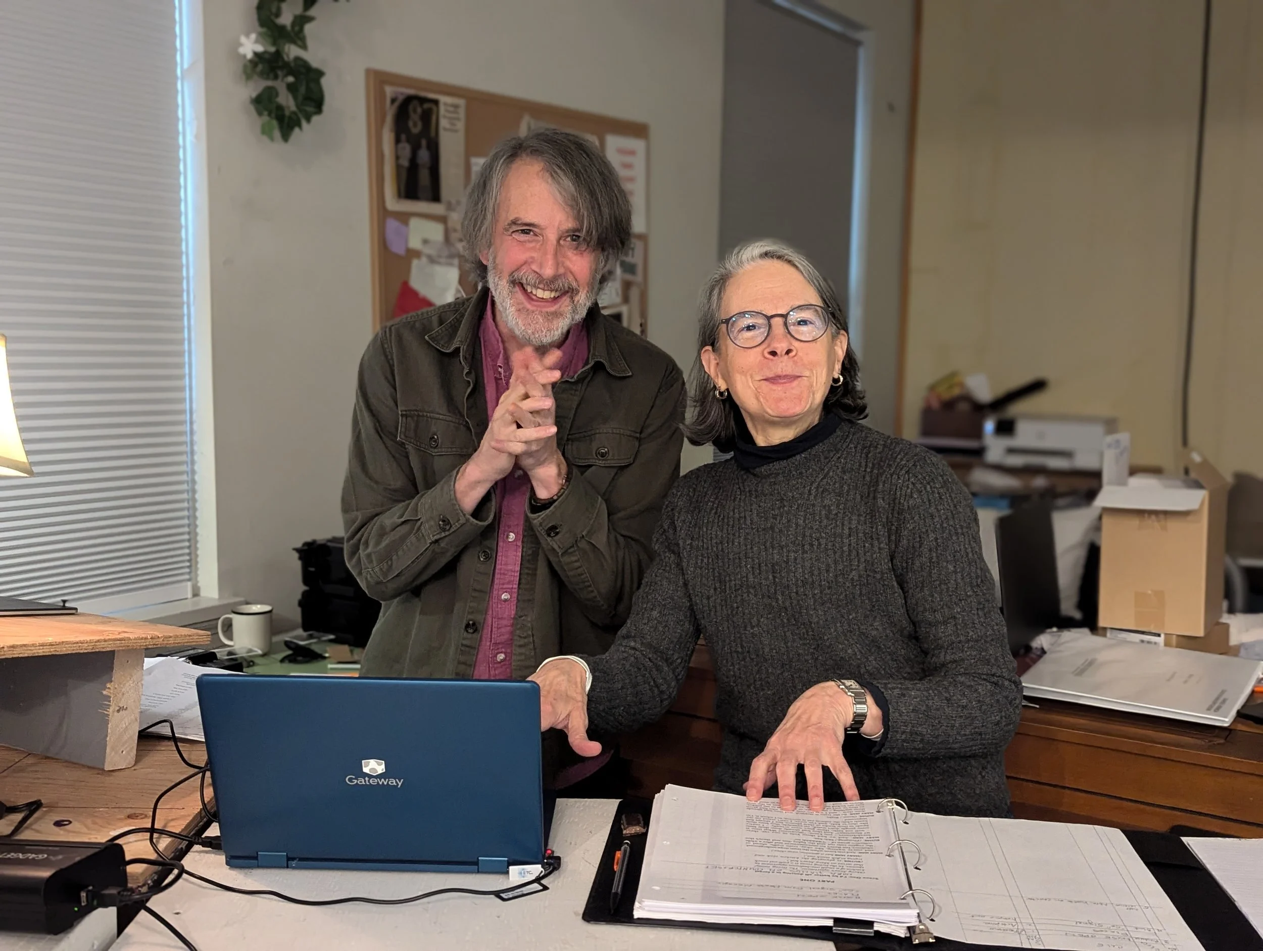 A man and woman in an office, smiling and shaking hands over a laptop with the brand Gateway, surrounded by papers, notebooks, and office supplies.