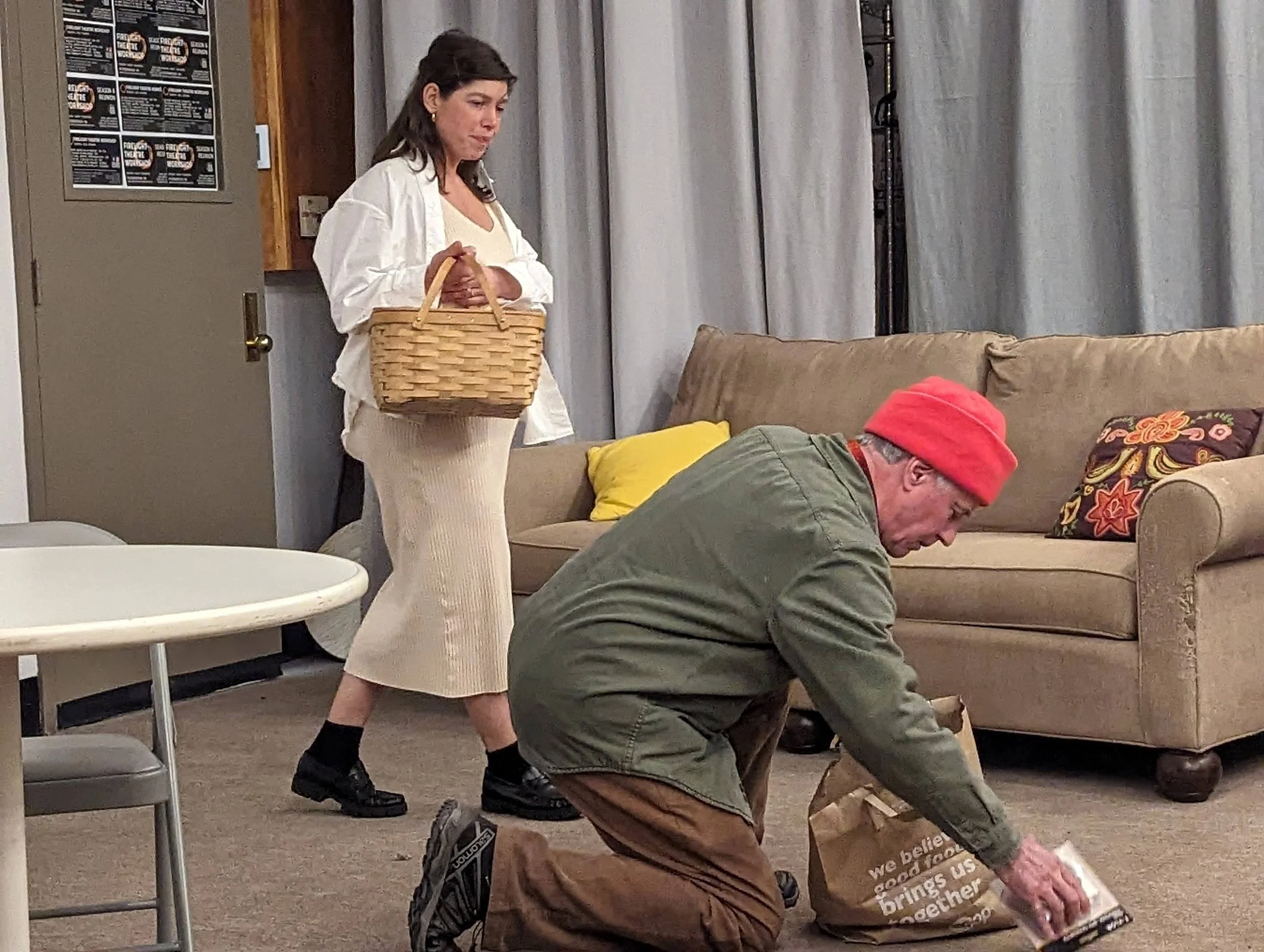A woman standing with a basket looks down at a man kneeling on the floor, selecting an item from a paper shopping bag.
