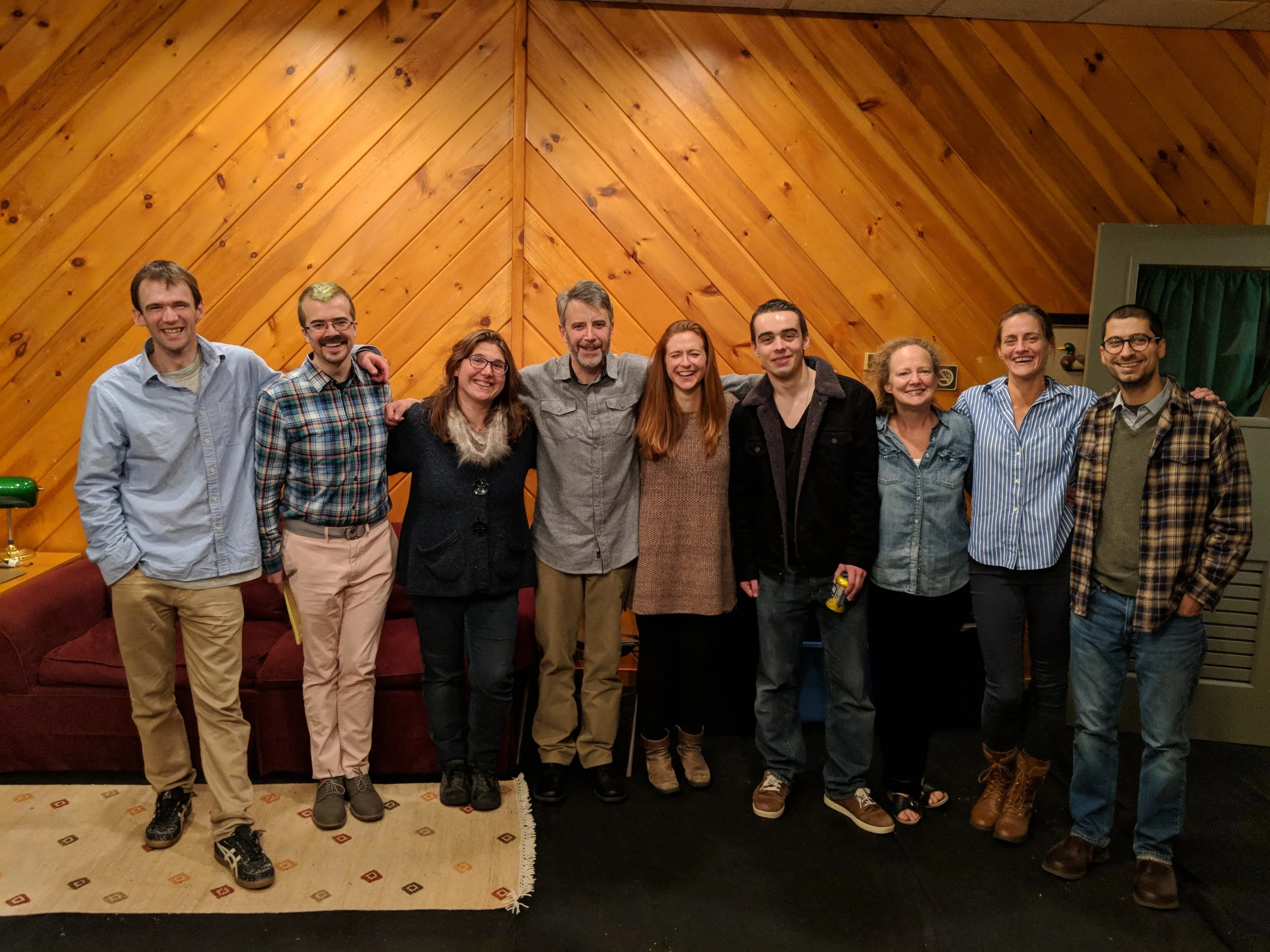 Nine people standing together in a room with wooden paneled walls, smiling for the photo. They have their arms around each other, indicating friendship or camaraderie.