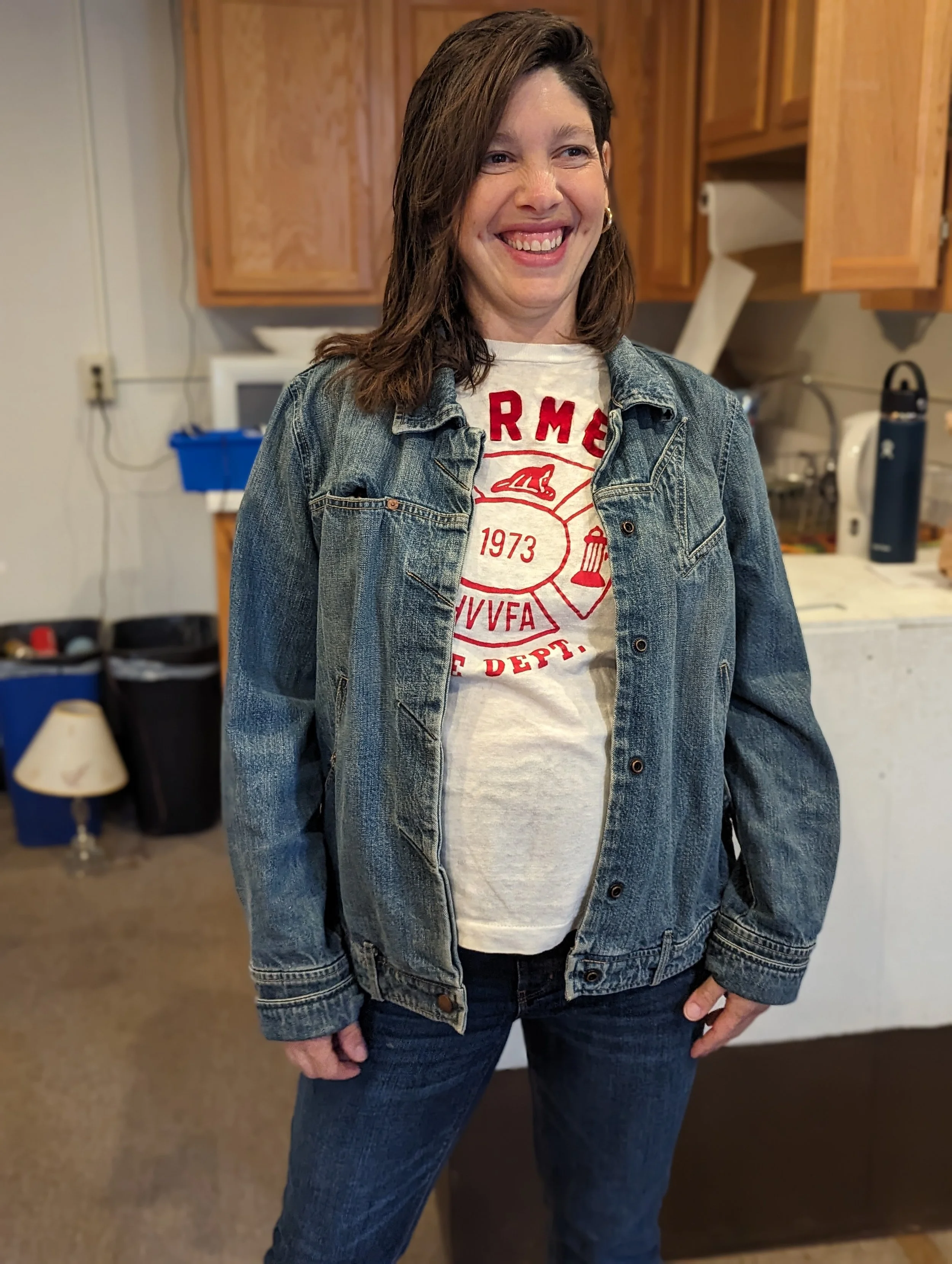A woman with shoulder-length brown hair, wearing a denim jacket over a white T-shirt with red print. She is smiling and standing in a kitchen with wooden cabinets and various appliances and items in the background.