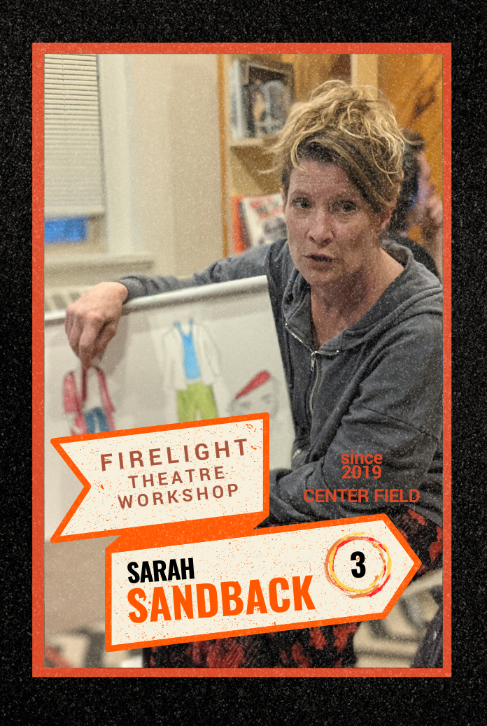 A woman with short blonde hair and a gray hoodie sitting in a classroom or workshop setting, holding a sign about a Firelight Theatre Workshop. The sign includes her name, Sarah Sandback, and her role as an artistic associate.