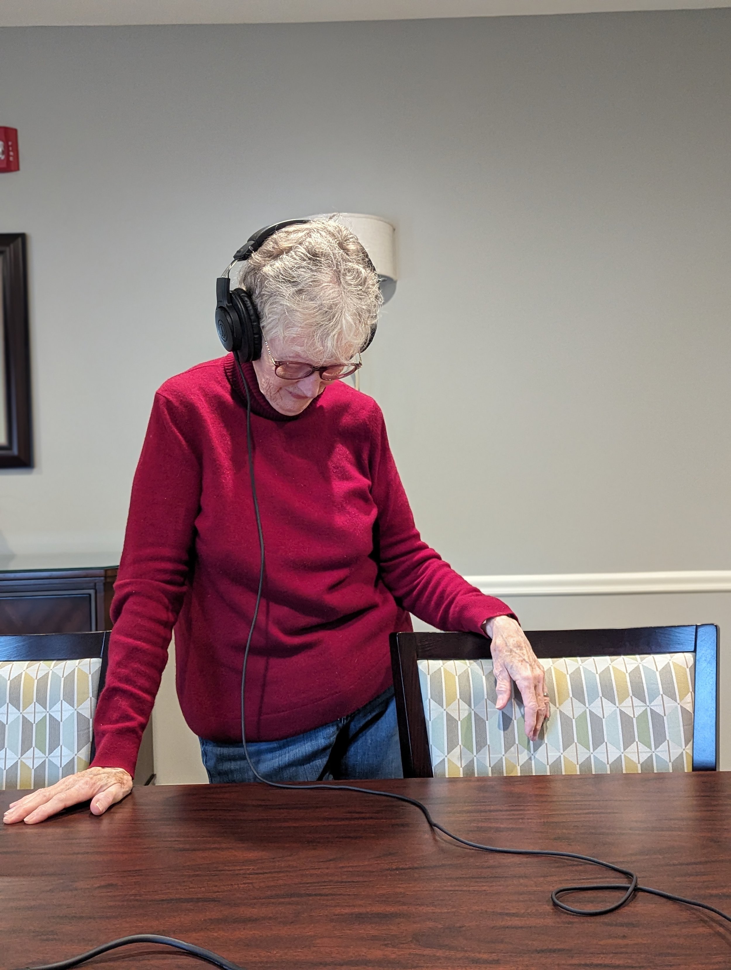 An elderly woman with gray curly hair and glasses is wearing large black headphones and standing by a wooden dining table. She is wearing a red sweater and blue jeans, with her right hand resting on the chair and her left hand on the table. The backg