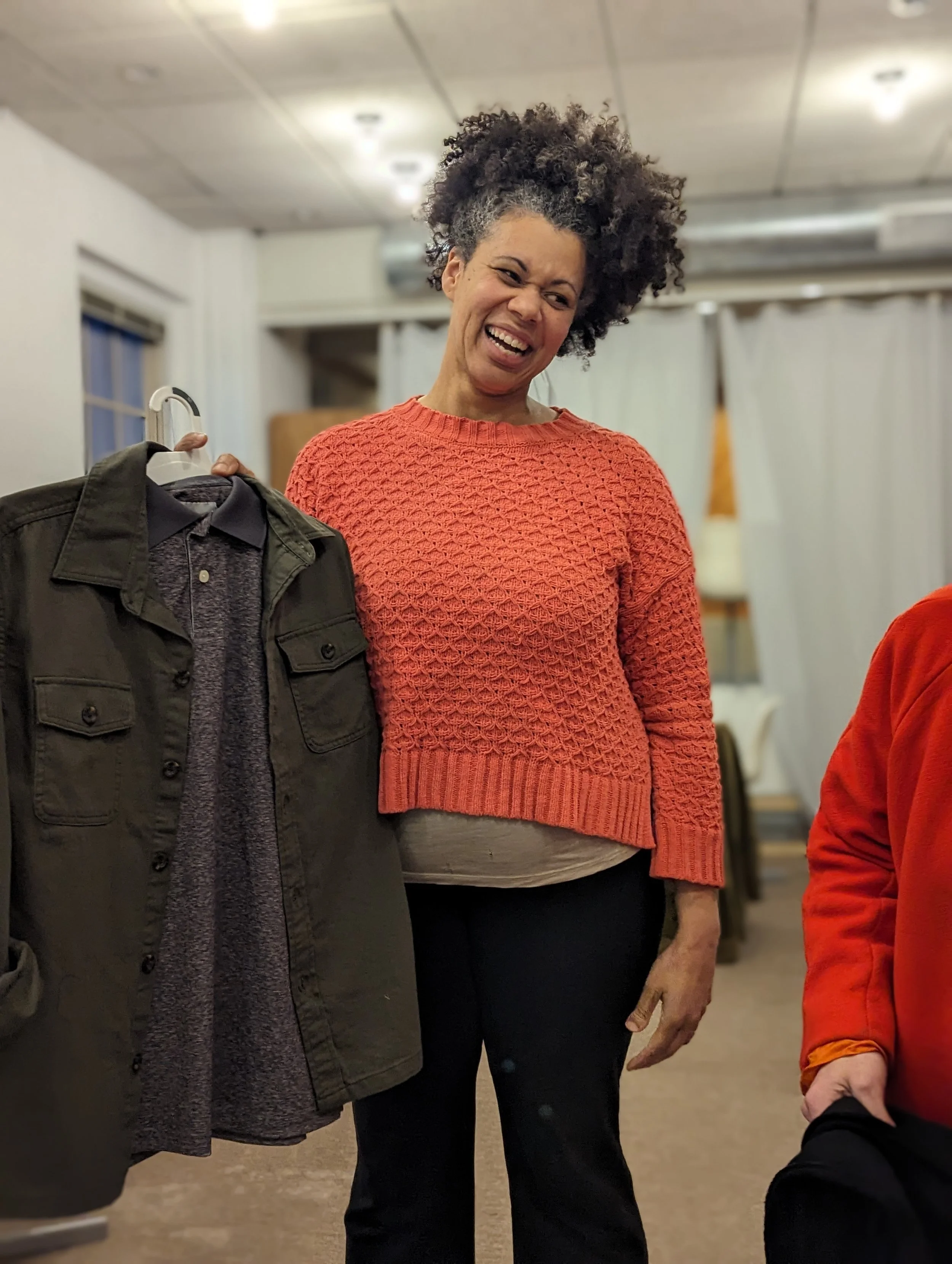 Woman with curly hair wearing a red textured sweater, standing indoors near a clothing rack with a jacket and shirt, smiling.