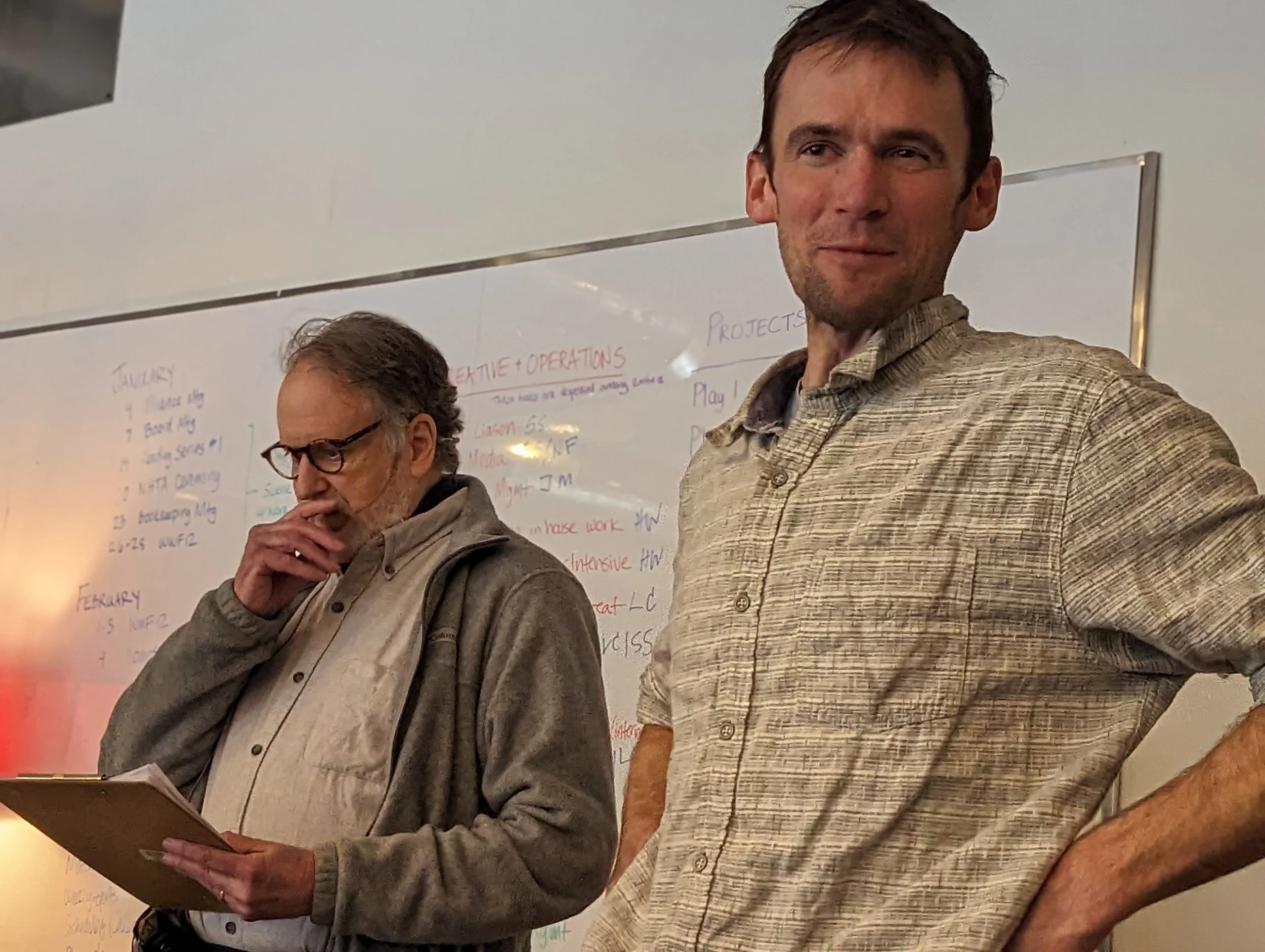 Two men standing in front of a whiteboard, one older with glasses and gray hair looking at notes, the other younger with short brown hair looking forward.