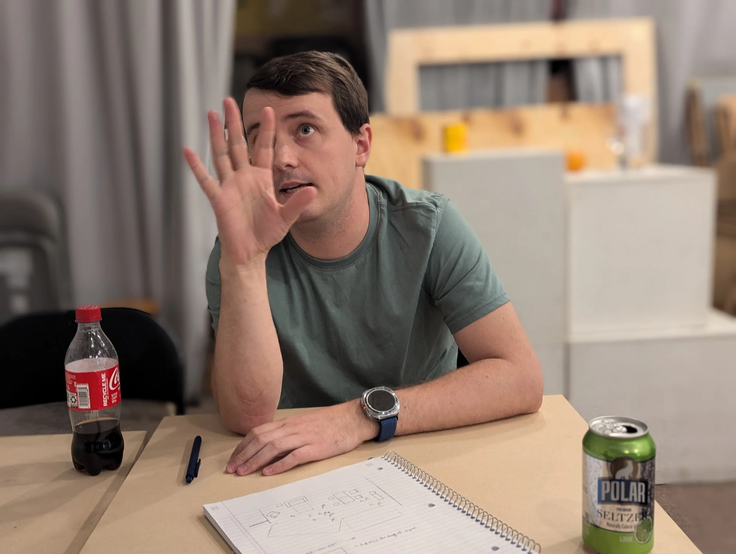A man with short dark hair wearing a green T-shirt and a watch on his wrist, sitting at a table with a Coca-Cola bottle, a notebook with sketches, a pen, and a can of Polar Seltzer. He is gesturing with his hand, pressing it near his face and looking