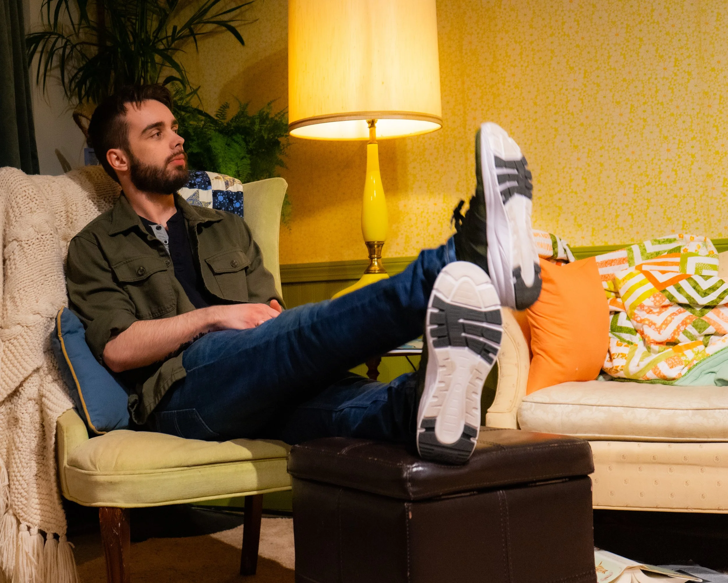 A young man with a beard sitting on a yellow couch, wearing a green jacket and blue jeans, with his legs extended and resting on a dark brown ottoman. The living room has a yellow patterned wall, a yellow lampshade, a green side table, and colorful p