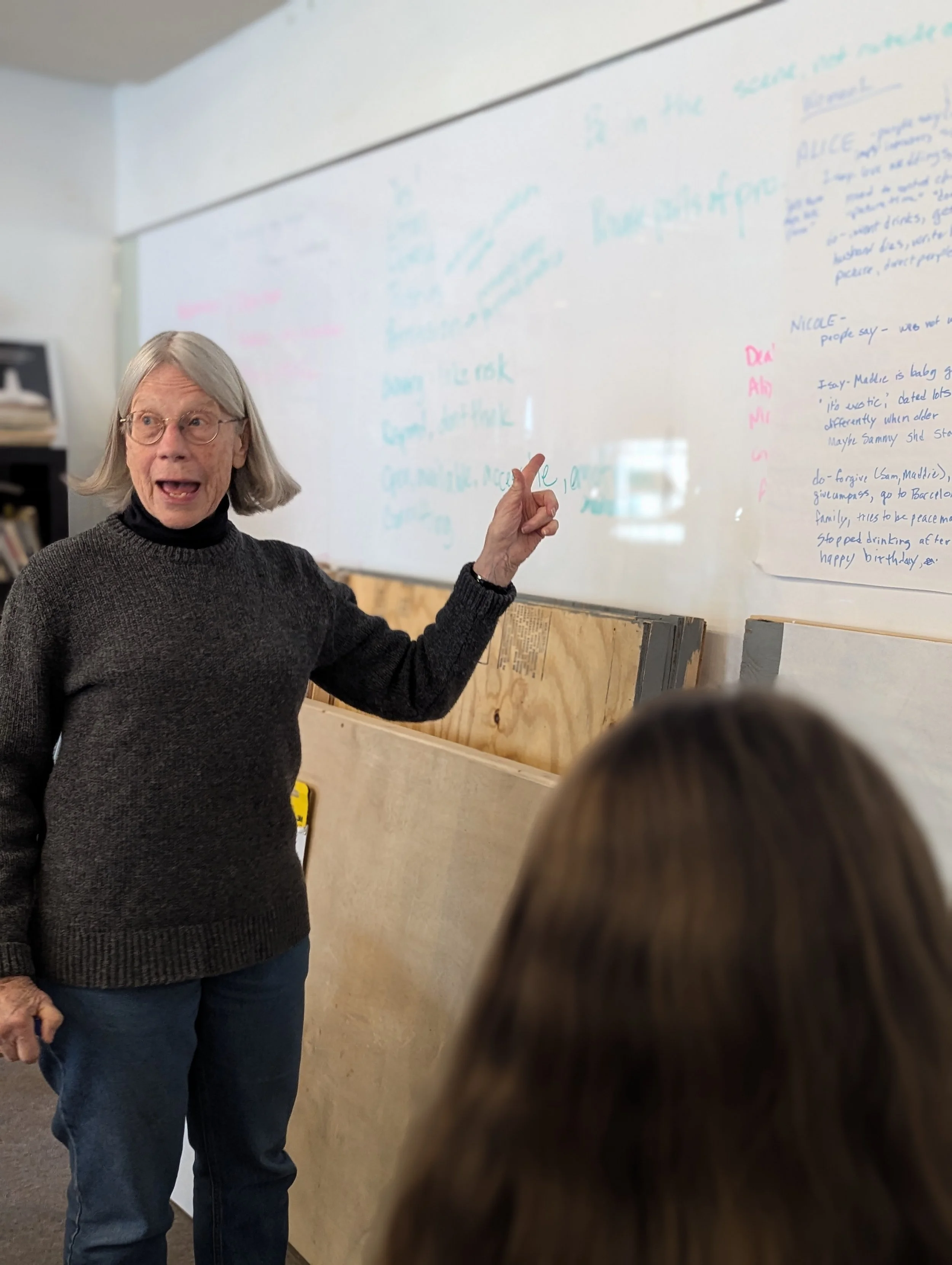 An elderly woman with gray hair, glasses, wearing a dark gray sweater, is pointing at a whiteboard with colorful writing on it. She appears to be teaching or giving a presentation to a person with long brown hair, who is partially visible in the fore