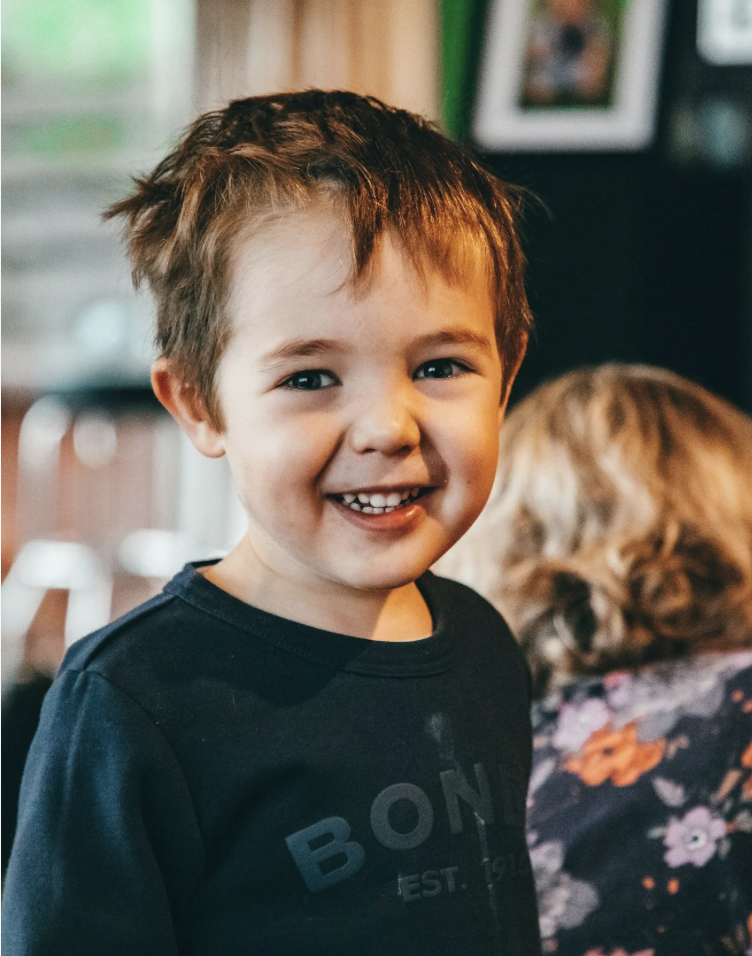 Smiling young boy with short brown hair, wearing a black shirt, indoors with blurred background of a woman and picture frames. Happy Face. What Empowered SLT aims to support children with by becoming confident communicators.
