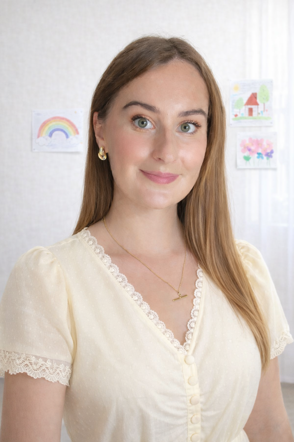 Meet Emma. A friendly woman with long, light brown hair, standing indoors, surrounded by children's drawings of a rainbow, a house, and flowers on the wall in the background. Friendly face of Empowered Speech & Language Therapy