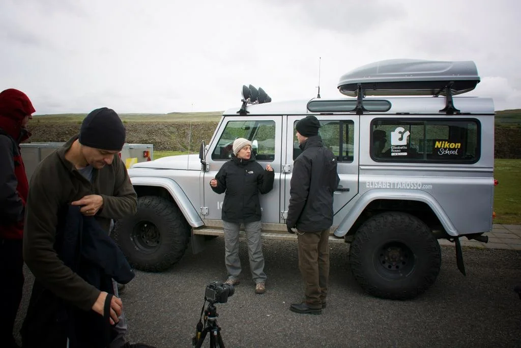 Quattro persone con abbigliamento invernale sono accanto a un veicolo fuoristrada grigio con attrezzature sulla parte superiore, in un paesaggio aperto sotto un cielo nuvoloso, mentre una di loro parla e le altre preparano l'attrezzatura.