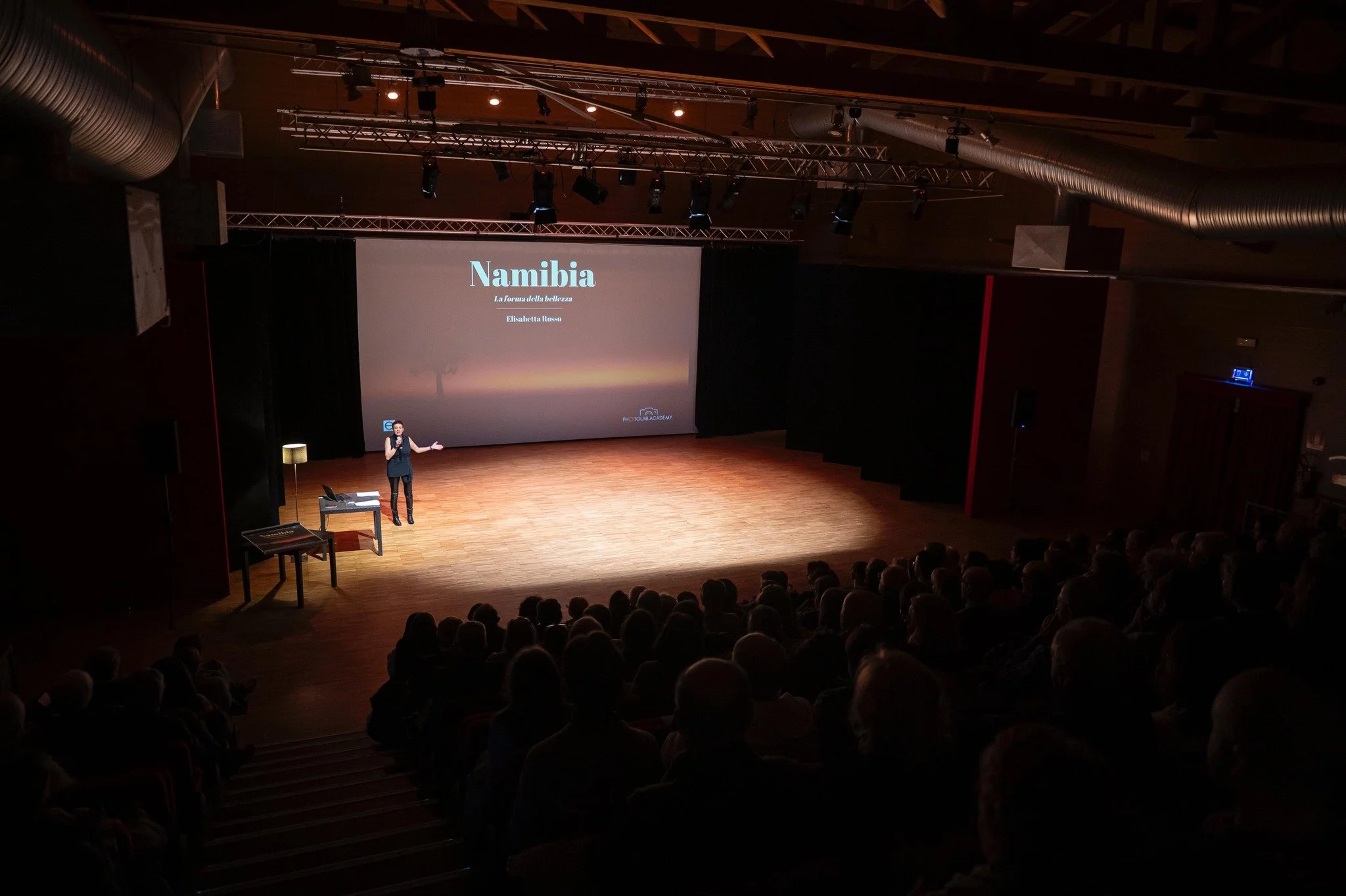 Un'oratrice sta facendo una presentazione su un palco in un teatro di fronte a un pubblico numeroso. Sullo schermo si legge 'Namibia La forma della bellezza Elisa Resso'.