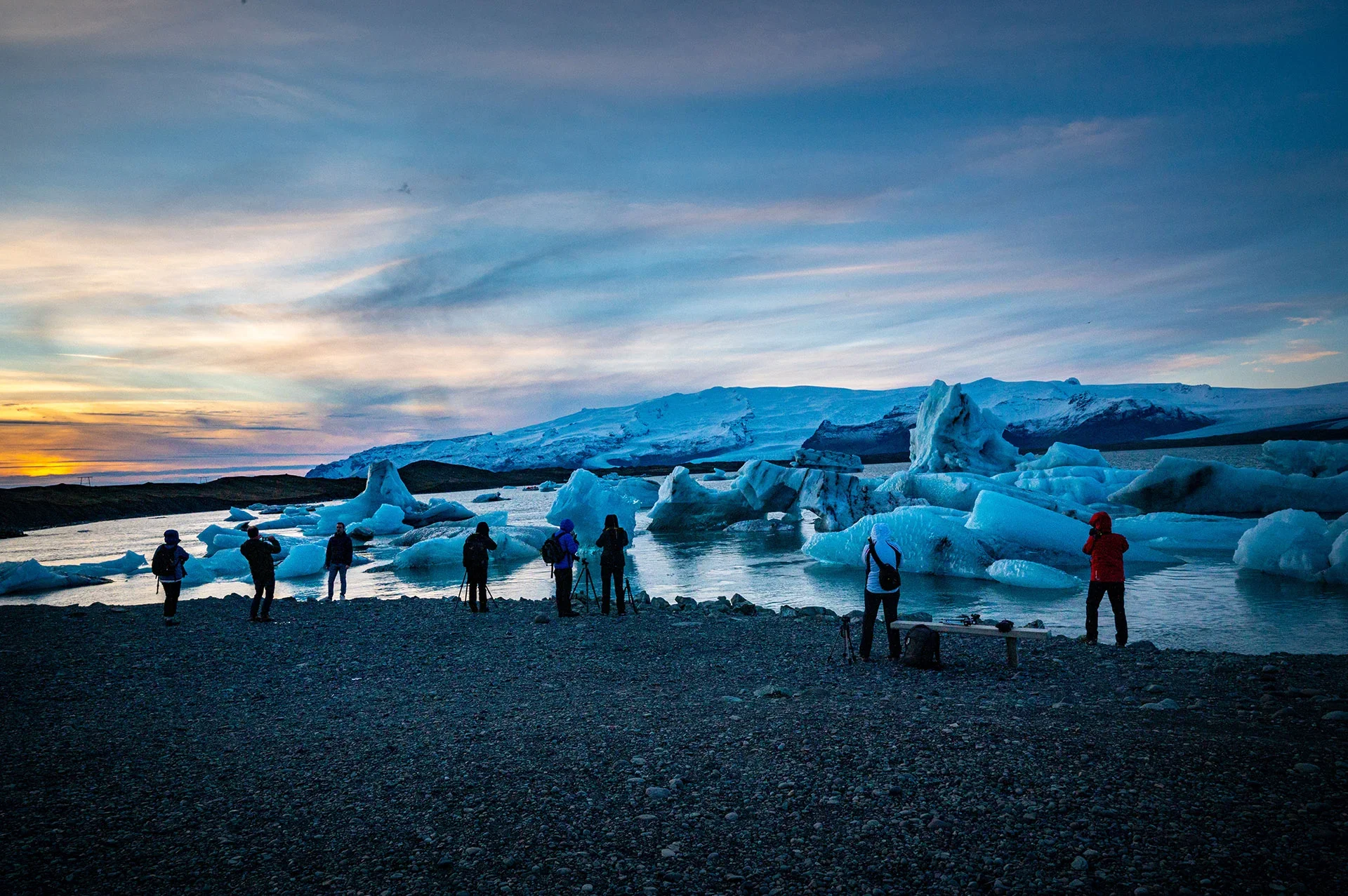 Gruppo di persone sulla riva di un lago con iceberg e montagne sullo sfondo, al tramonto.