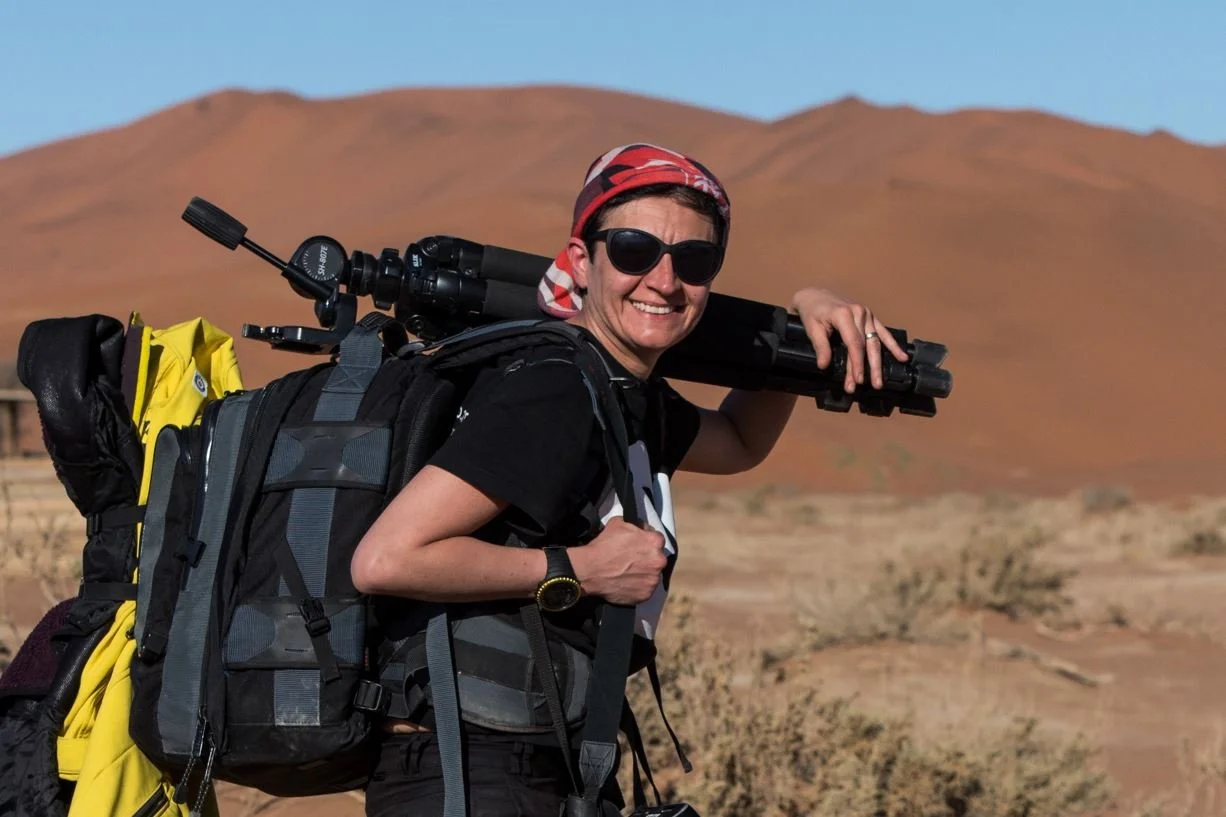 Donna sorridente con occhiali scuri e bandana sulla testa, in un deserto con montagne sullo sfondo, che porta uno zaino grande e una fotocamera con treppiede sulle spalle.