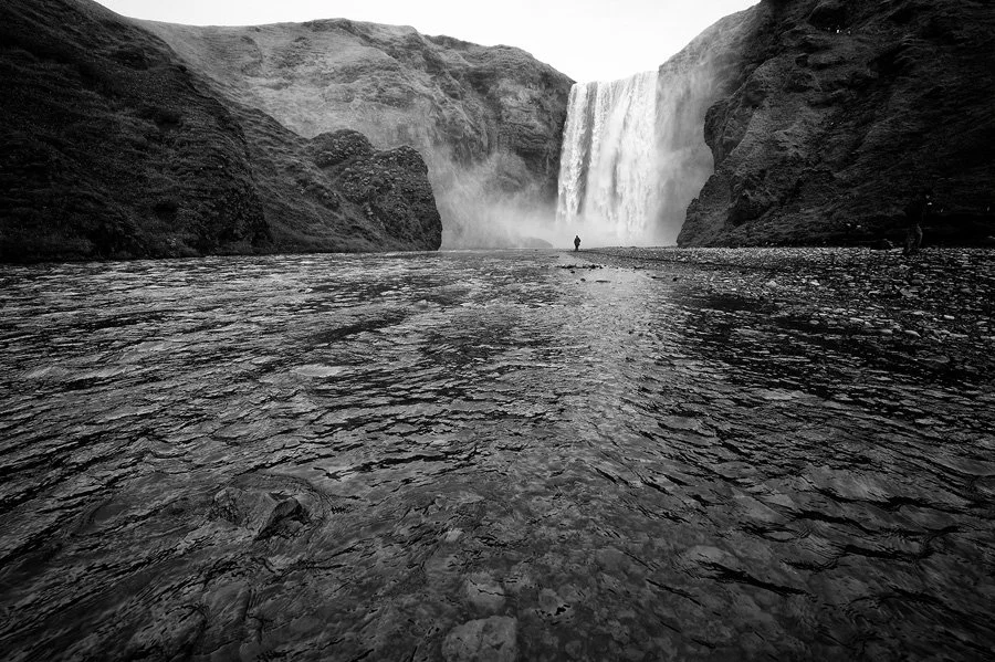 Cascata in un paesaggio naturale, con acqua che scorre e una persona in lontananza, in bianco e nero.