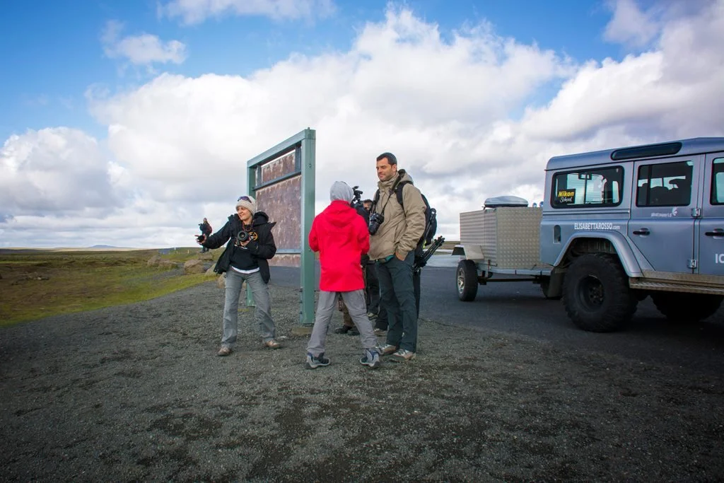 Gruppo di persone con attrezzature fotografiche accanto a un veicolo overland in un paesaggio aperto con nuvole nel cielo.