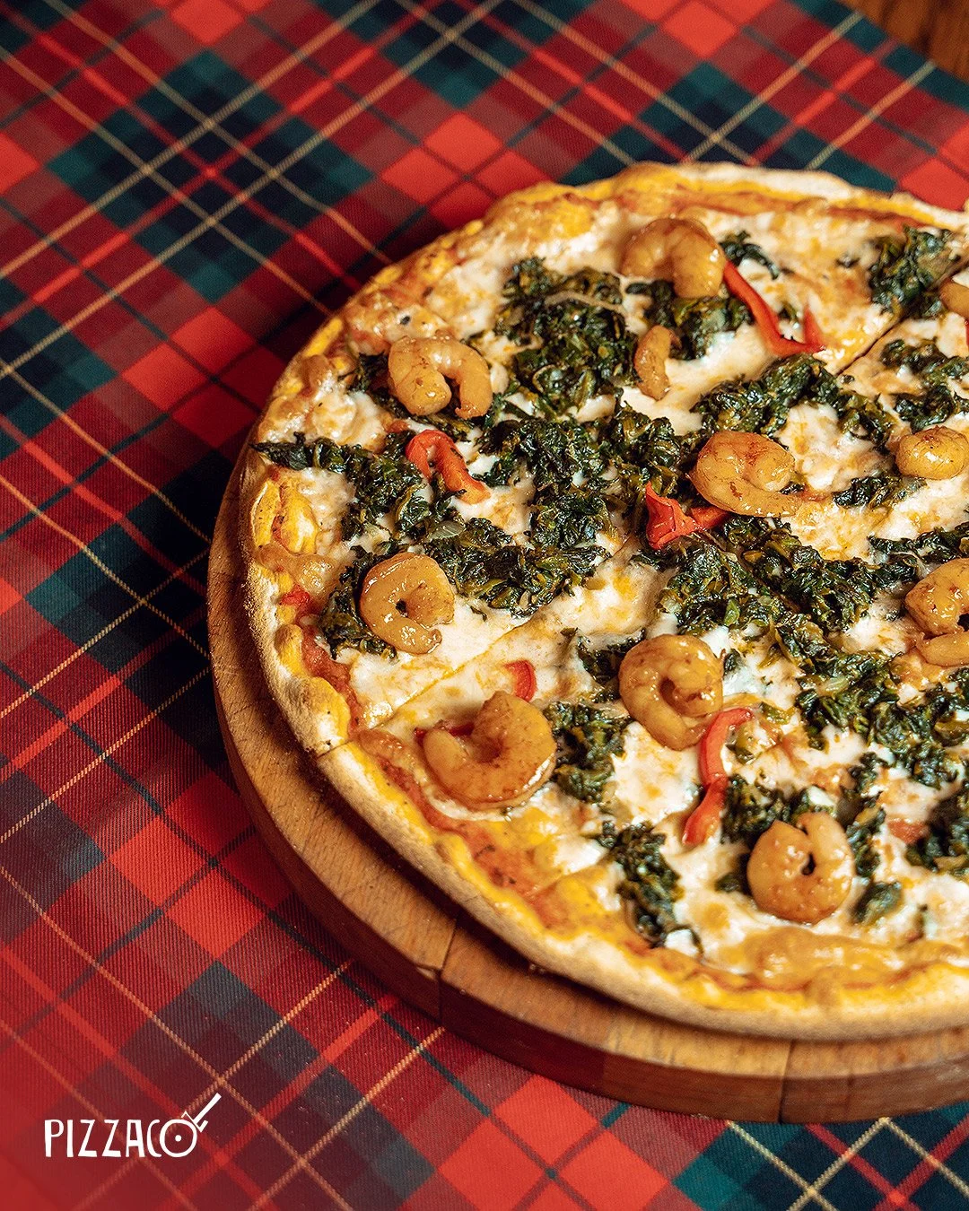 Pizzanın üzeri karides, ıspanak ve kırmızı biber ile süslenmiş, yuvarlak ve odun fırını tarzı tahtadan servis tabağında sunulmuş. Altında kırmızı ve yeşil kontrol deseni olan masa örtüsü var.