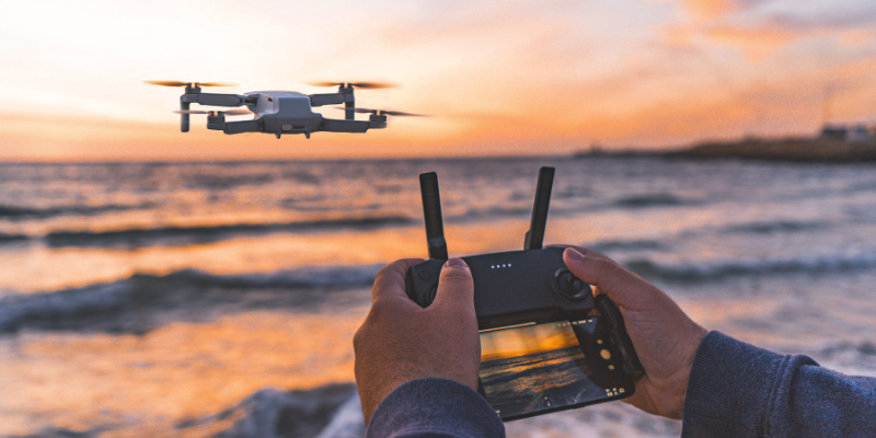 Bir kişi, deniz kıyısında akşam gün batımında, drone kontrol cihazıyla drone'u havada uçuruyor.