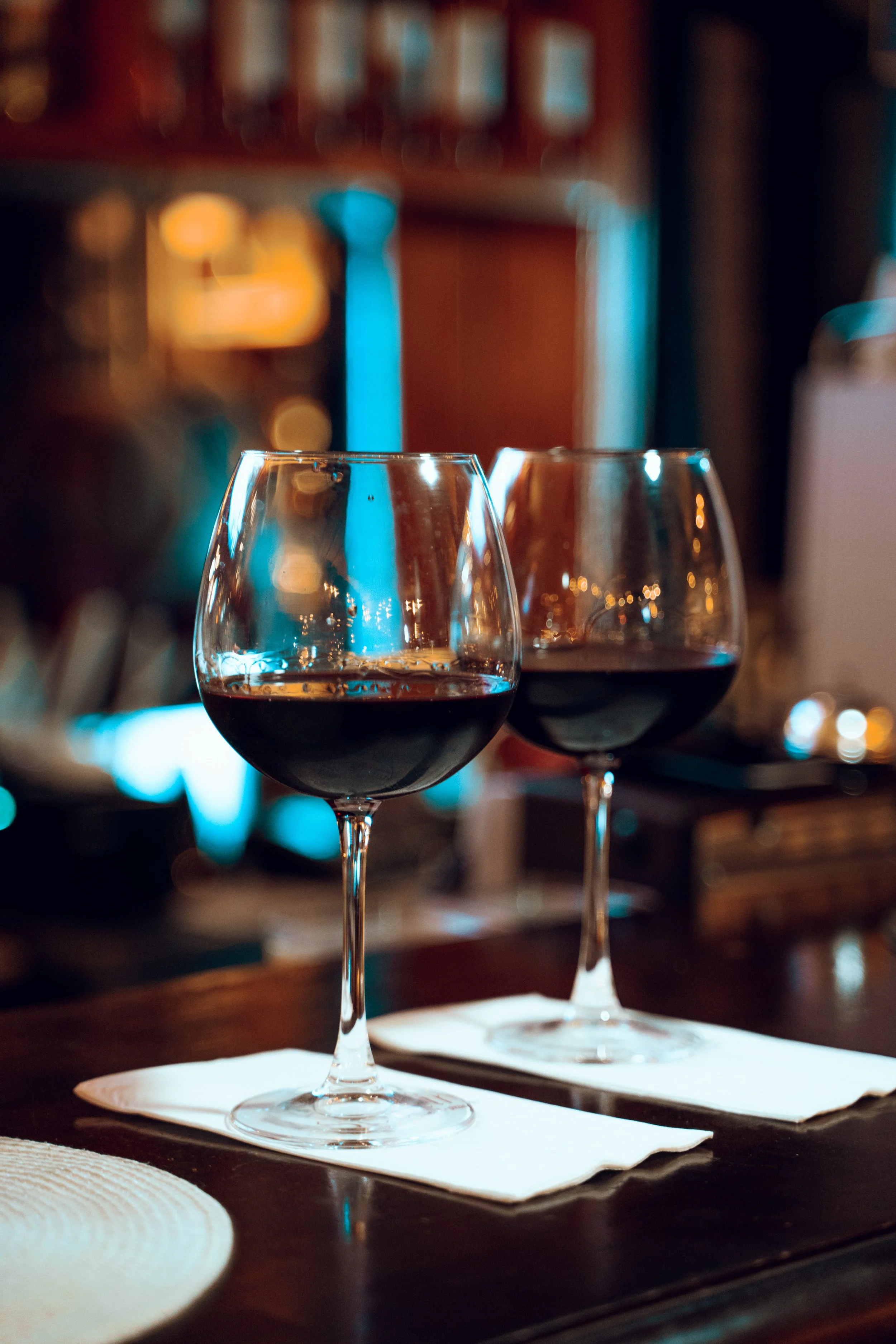 İki adet kırmızı şarap kadehi, içlerinde kırmızı şarap ile, masada yan yana duruyor. Lafsız altlıklar üzerinde yer alıyorlar, arka planda ise sıcak ve loş bir iç mekan var.