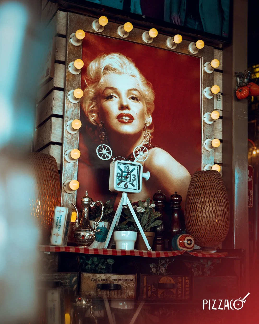 İç mekan dekorasyonu, büyük bir Marilyn Monroe portresi ve etrafında ışıklandırmalar, masaüstü saat, bitkiler ve çeşitli dekoratif nesneler ile Pizzaco logosu.