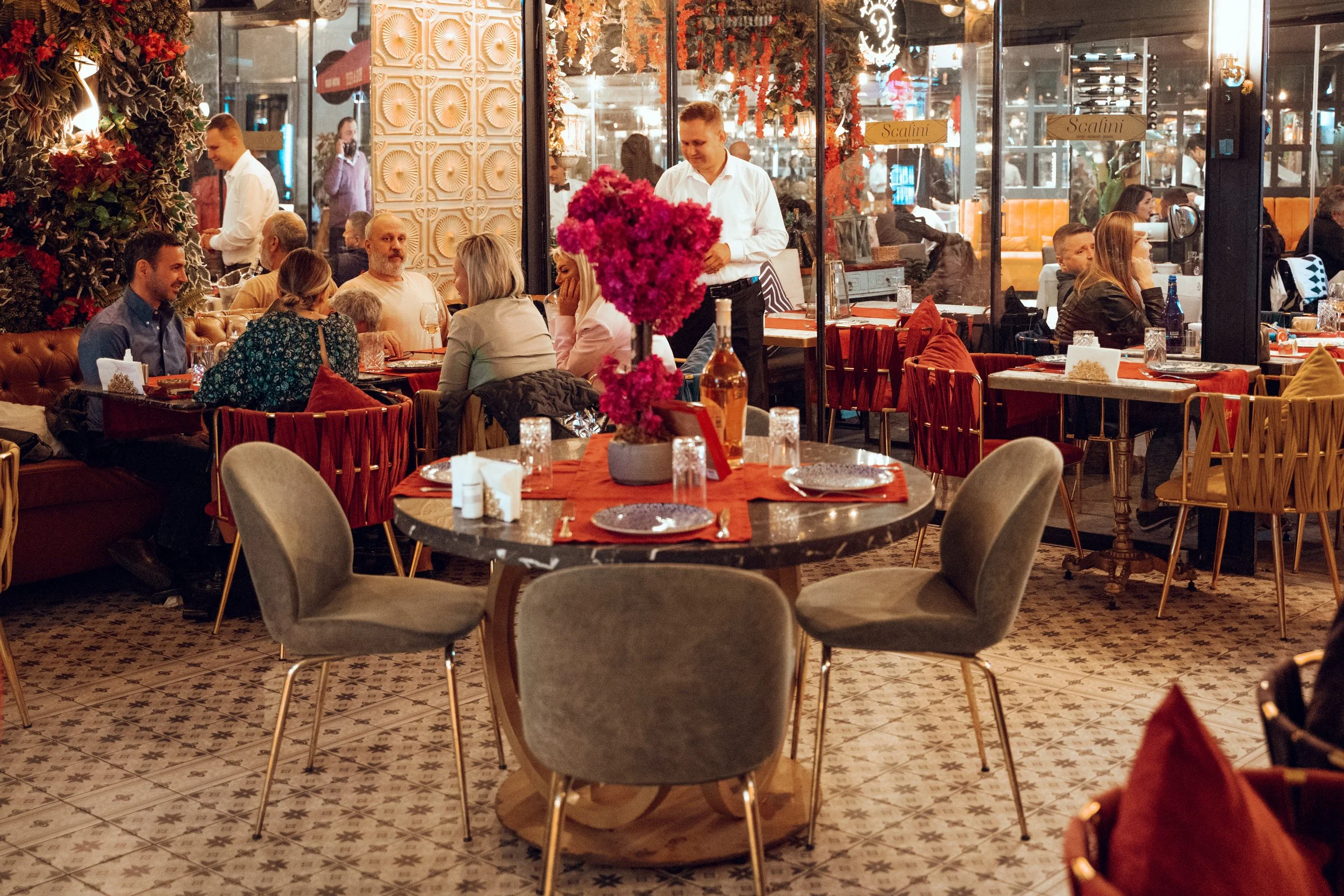 Bir restoran veya kafe içi, ortasında büyük yuvarlak masada pembe çiçeklerle süslenmiş, üzerinde birkaç tabak ve bardak bulunan dekoratif bir çiçek düzeni. Arka planda birkaç masa ve sandalye, müşteriler ve birkaç garson görülüyor. Mekanın dekorasyon