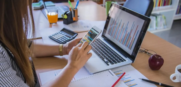 Kişi masa üzerinde laptop, telefon ve not defteri kullanarak çalışıyor, aynı zamanda telefonundan grafiklere bakıyor. Masada meyve, kalemler, cetvel ve kitaplar bulunuyor.