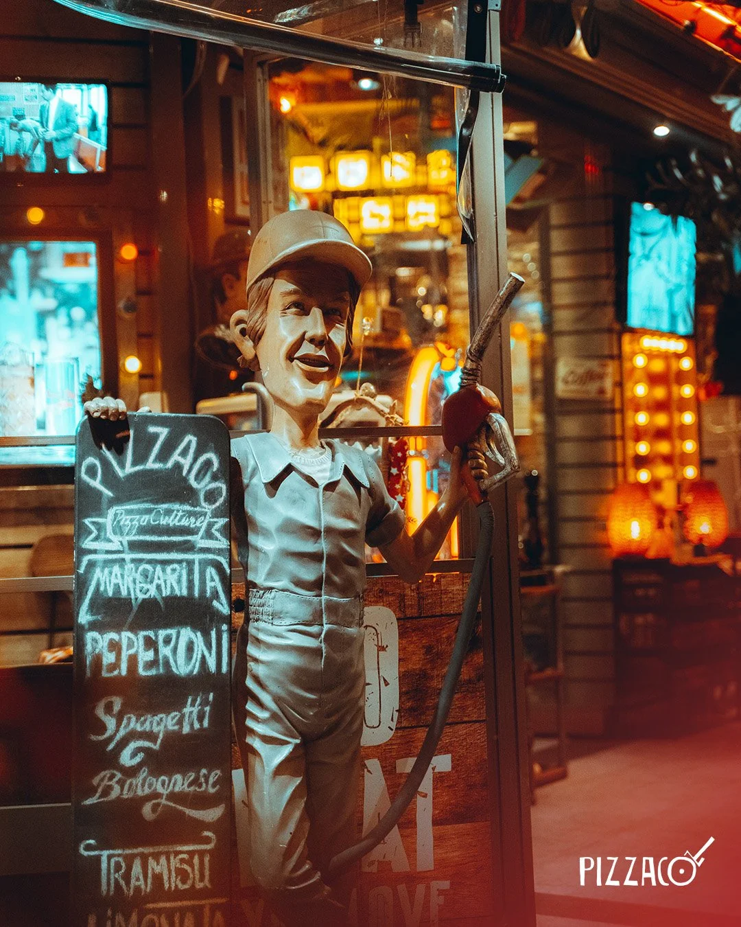Bir pizza restoranının önünde, şapka takmış, gülümseyen ve elinde büyük bir biber asili tutan bir figür. Arkada çeşitli ışıklı tabelalar ve iç mekân dekorasyonu görünuyor.