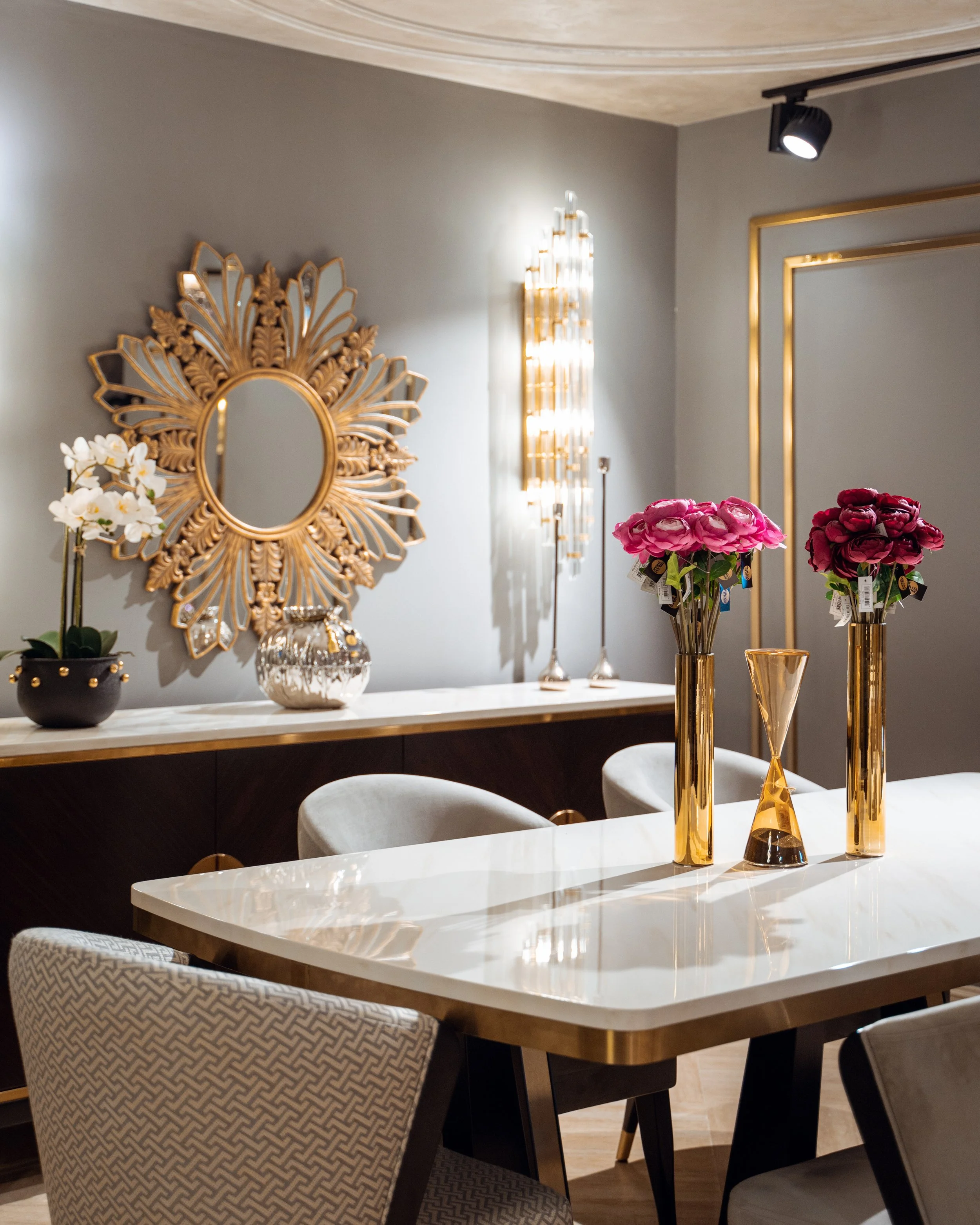 Şık bir dekoratif iç mekan görünümü. Duvarda altın rengi detaylı büyük bir ayna ve avize var. Masanın üzerinde altın detaylı vazo ve çiçekler bulunuyor.