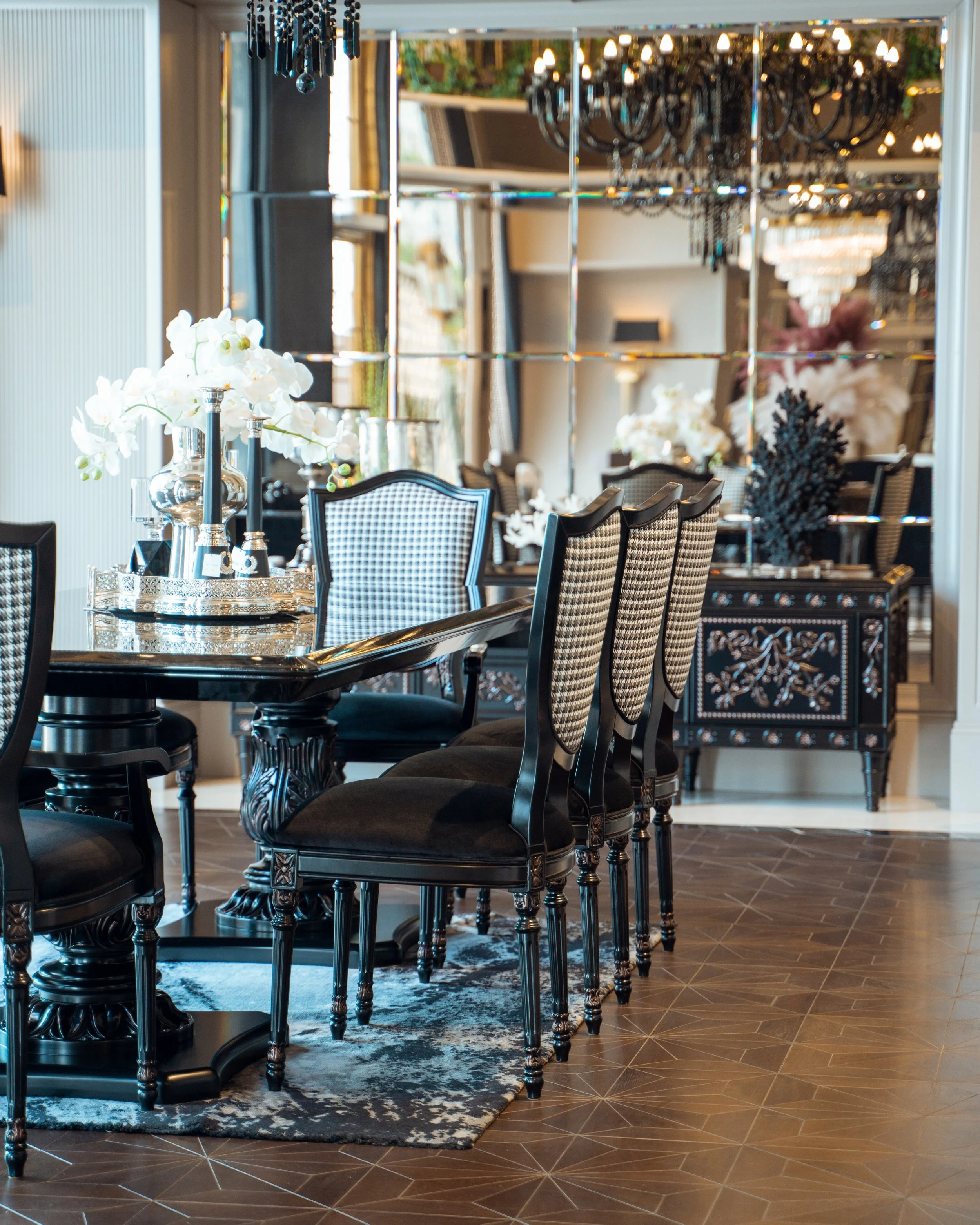 Lüks bir yemek odası, siyah ve beyaz desenli sandalyelerle ve büyük bir masa, ortasında beyaz çiçeklerle süslenmiş dekoratif devamlılar. Arka planda büyük aynalar ve kristal avizeler bulunuyor.