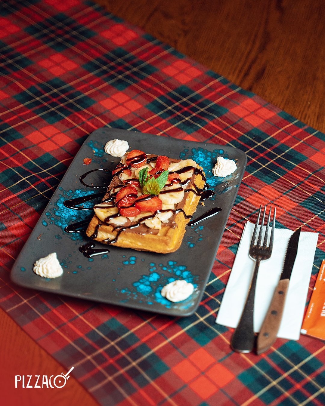 Çikolatalı ve meyveli waffle, üzerinde çilek, muz ve frambuaz dilimleri ile yeşil nane yaprağı bulunan tatlı. Yanında küçük kremalar ve çikolata sosu ile servis edilmiş, arka planda kırmızı, yeşil ve lacivert renkli ekose masa bezi üzerinde sunulmuş.