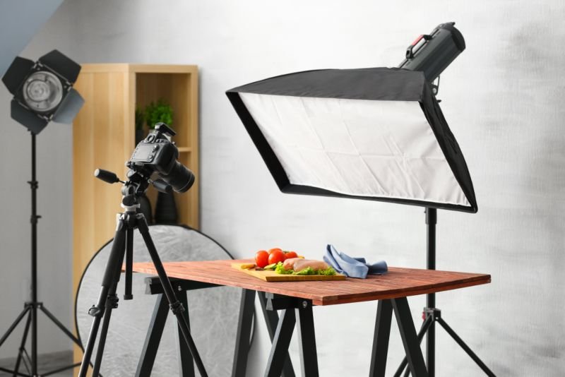 Bir mutfak veya stüdyo ortamında yemek fotoğrafı için hazırlık yapan bir video veya fotoğraf çekim sahnesi, kamera, ışıklandırma ekipmanları ve bir masa üzerine koyulmuş domatesler ve meyve sebze.