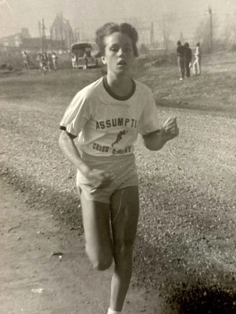 A young woman running outdoors on a gravel path, wearing a sports jersey with 'Assumption Cross Country' printed on it, in a vintage black-and-white photograph.
