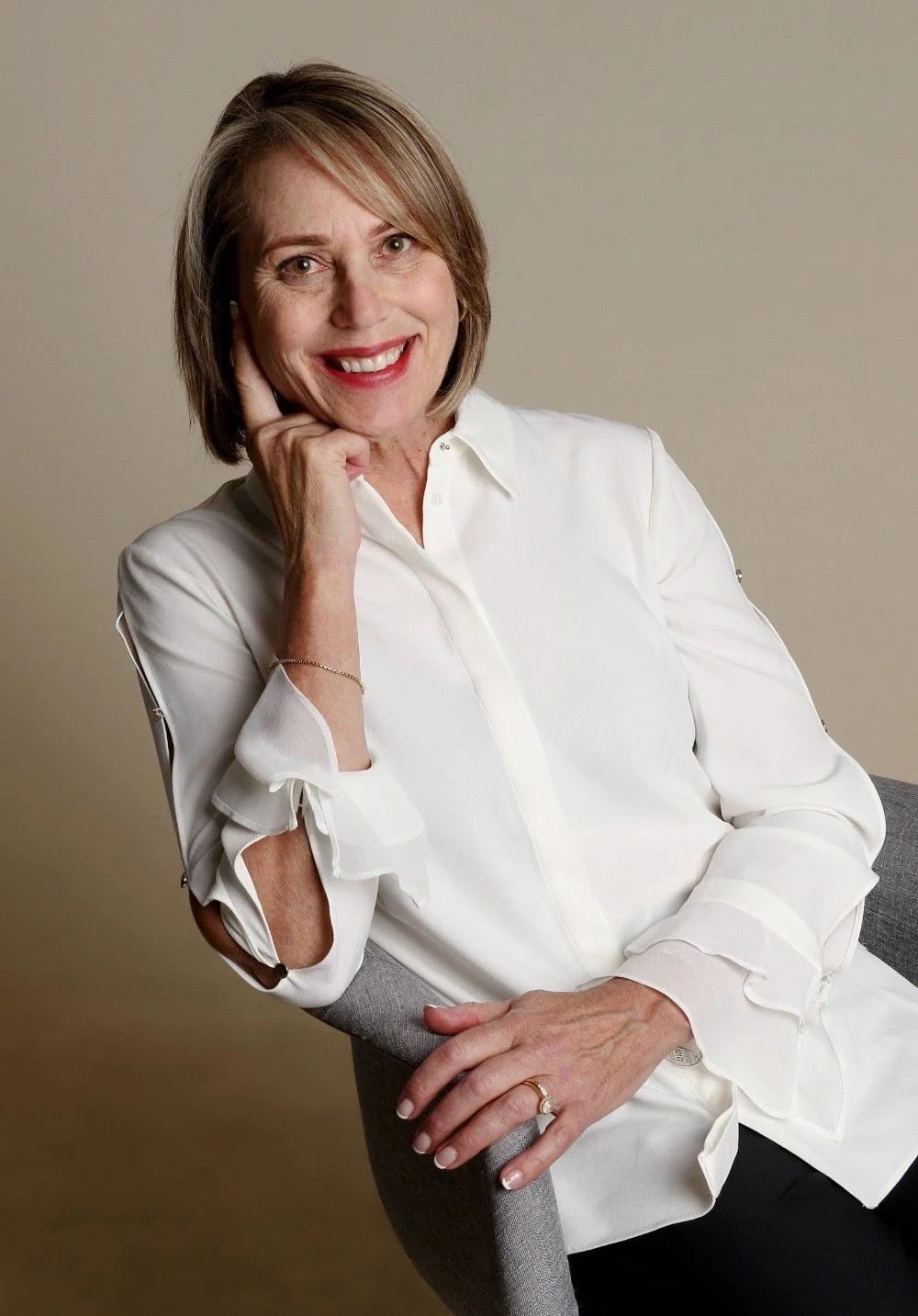A woman with shoulder-length blond hair, smiling, wearing a white blouse, sitting on a gray chair against a beige background.