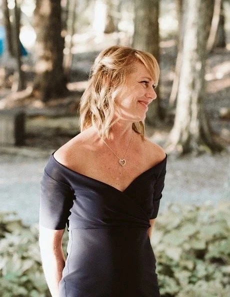 A woman with blonde hair wearing an off-the-shoulder black dress and a heart-shaped necklace, standing outdoors near trees and greenery.