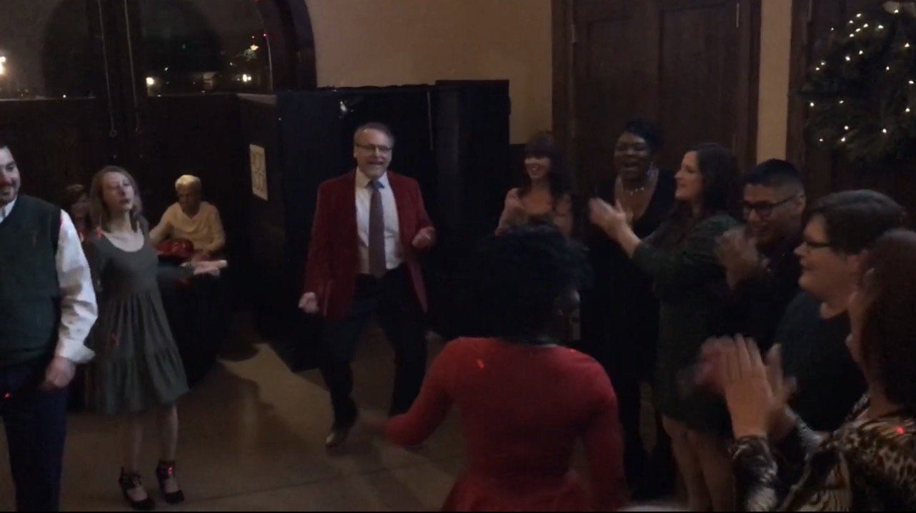 Group of people dancing and clapping in a warmly lit indoor setting, celebrating at a social gathering or party.