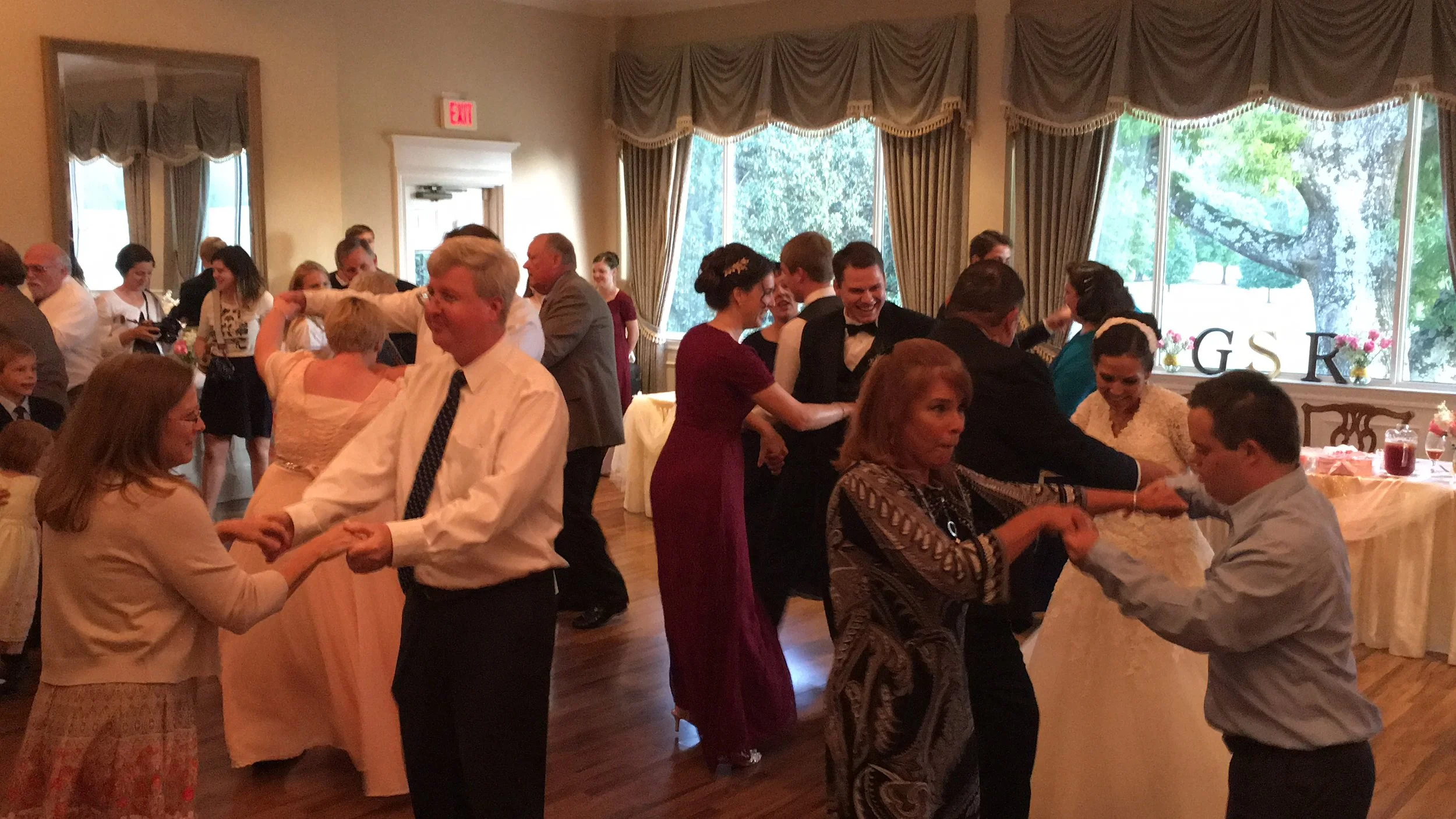 People dancing at a formal gathering in a decorated banquet hall with large windows and elegant curtains.