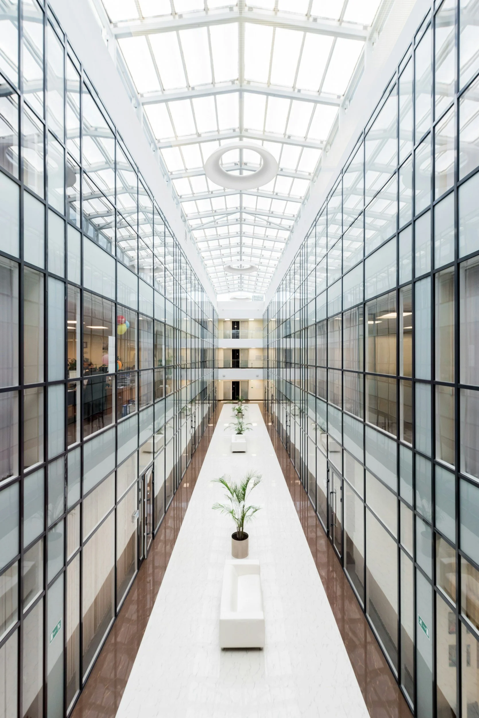 A modern multi-story office building atrium with glass walls, plants, white seating, and a glass ceiling.