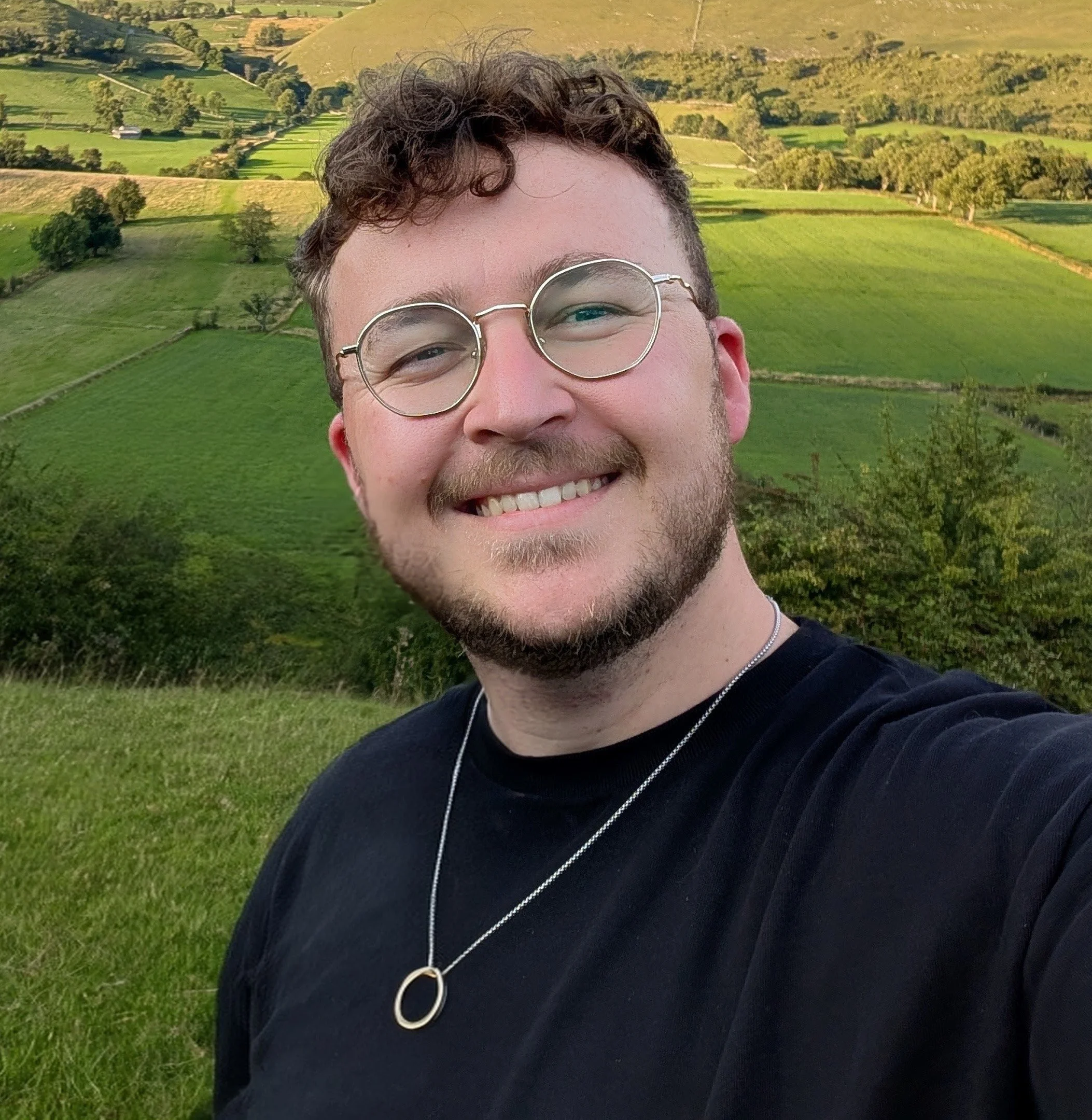 A smiling man with glasses and a necklace taking a selfie outdoors in front of rolling green hills and trees.