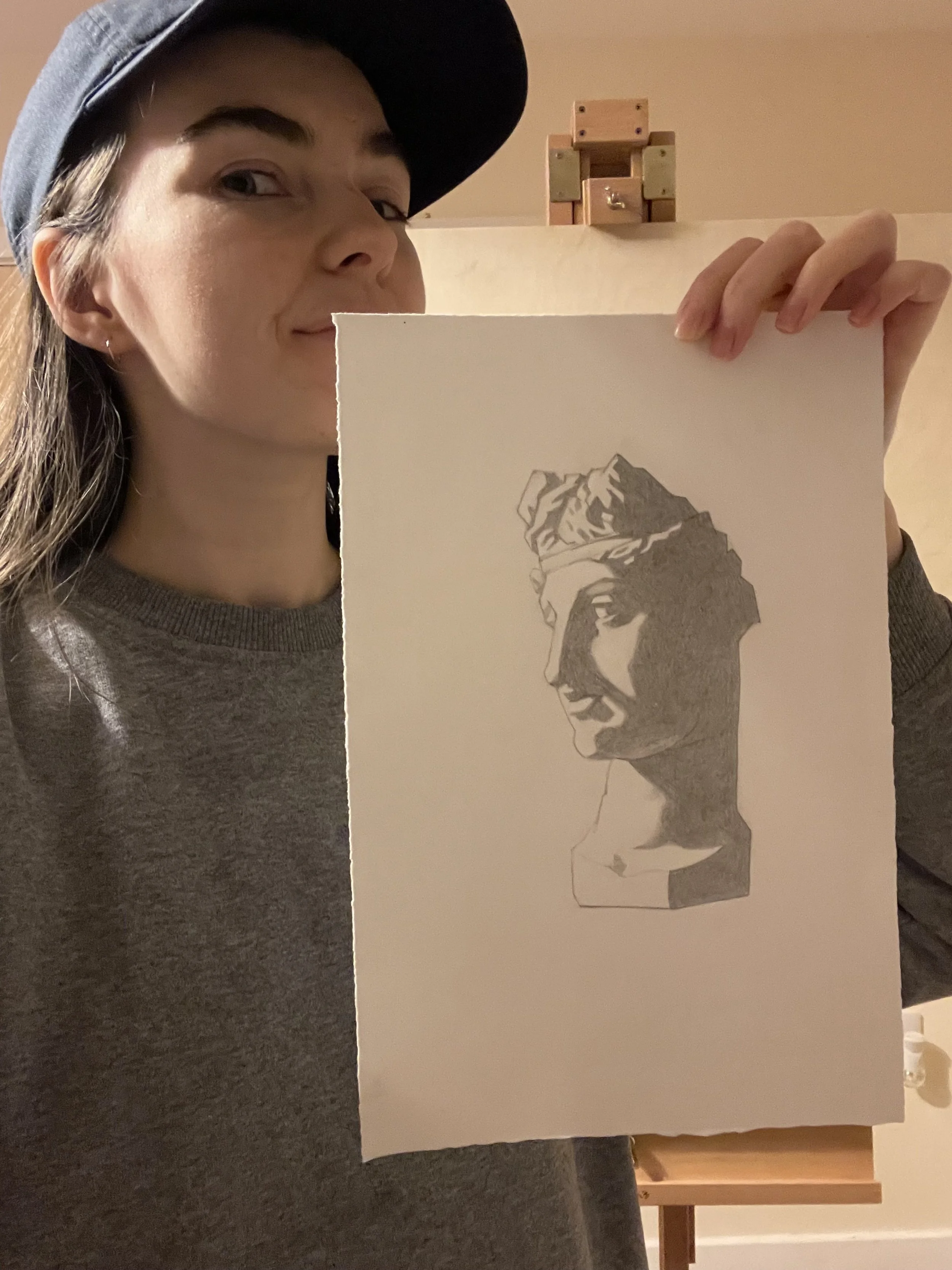 A woman wearing a gray shirt and black cap holding a drawing of a classical statue's bust. The drawing is on a piece of white paper with a gray shading sketch of the statue's head and face.